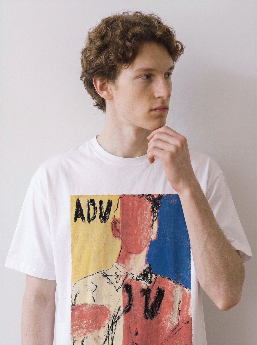 A male model resting his hand on his chin wearing a white abstract faceless portrait graphic tee, showcasing the vivid primary color blocks and indie art aesthetic.