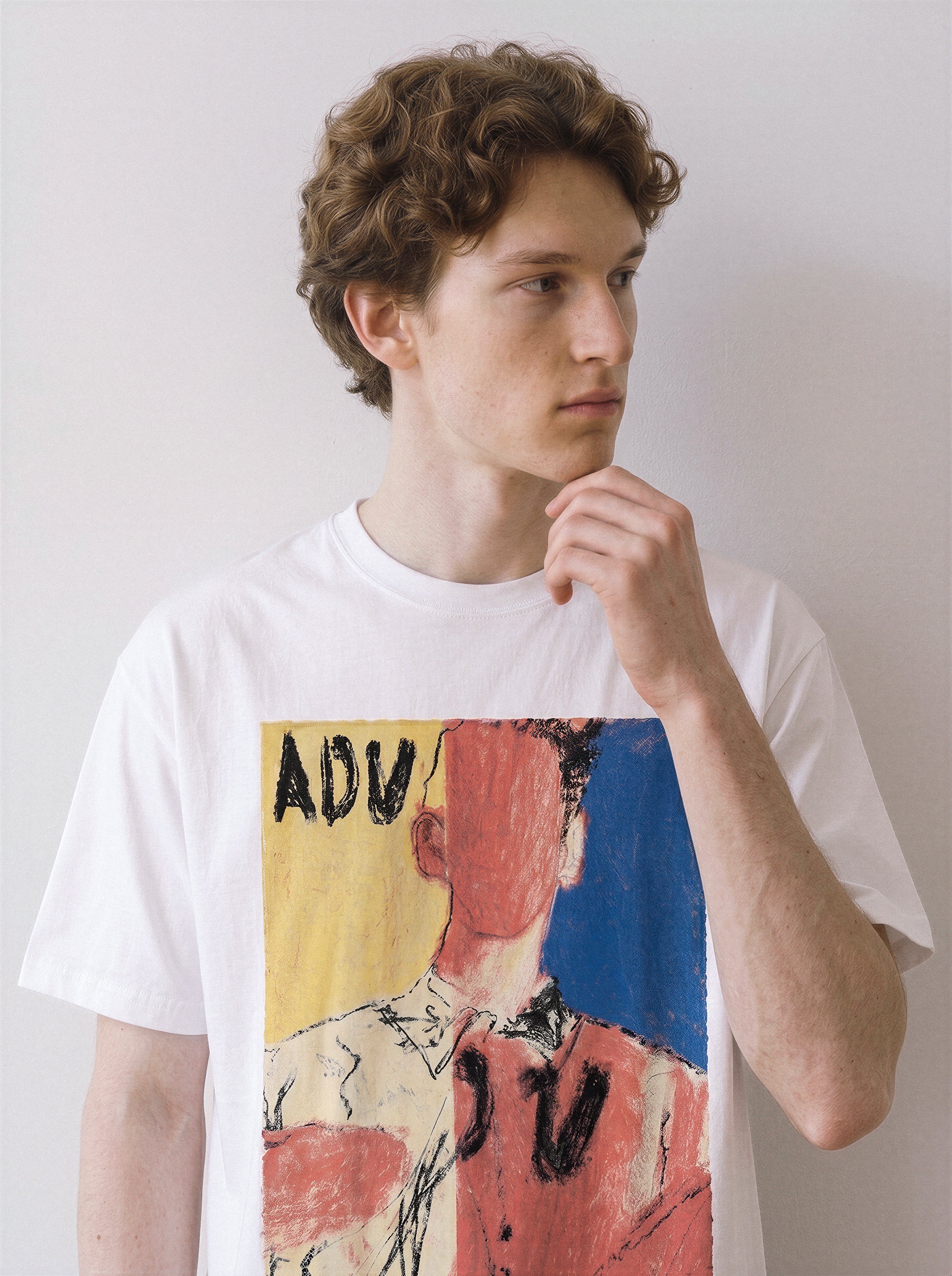 A male model resting his hand on his chin wearing a white abstract faceless portrait graphic tee, showcasing the vivid primary color blocks and indie art aesthetic.