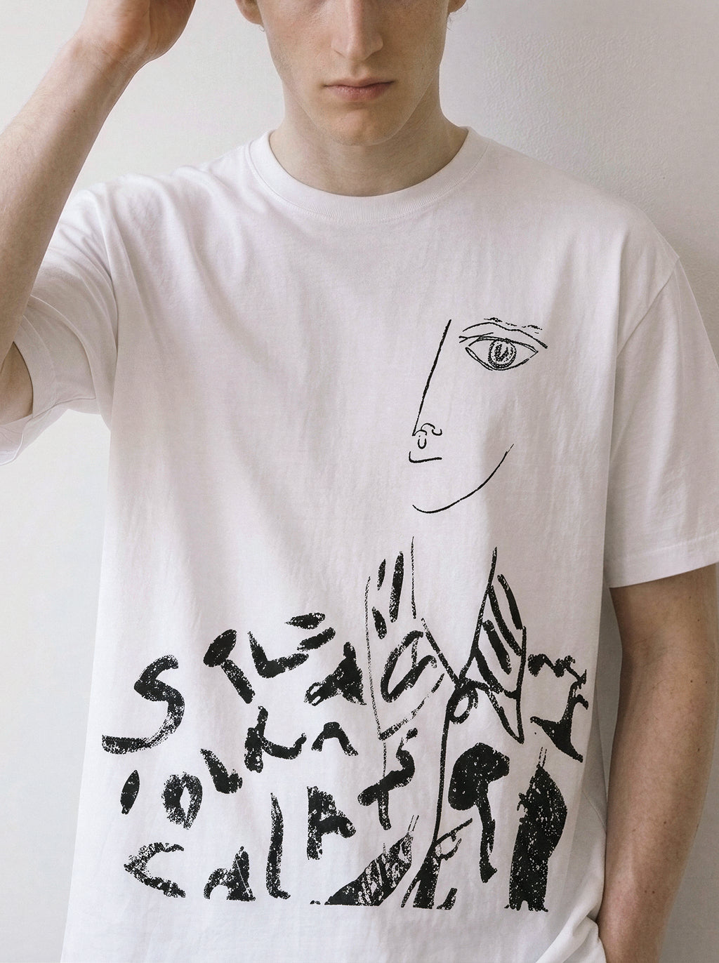 A white short-sleeve crewneck t-shirt featuring a black line art graphic of a face and abstract text, worn by a model against a plain light background.