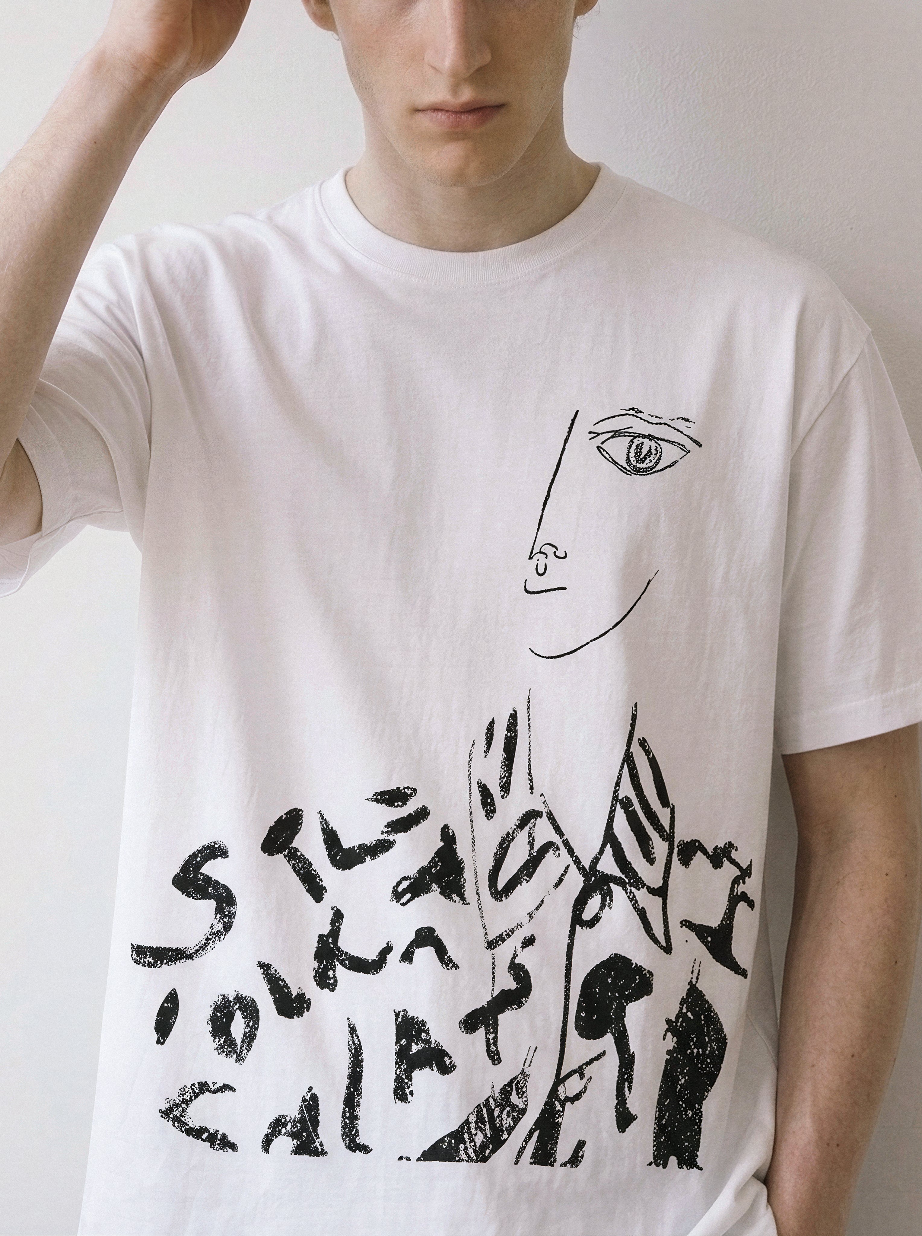 A white short-sleeve crewneck t-shirt featuring a black line art graphic of a face and abstract text, worn by a model against a plain light background.