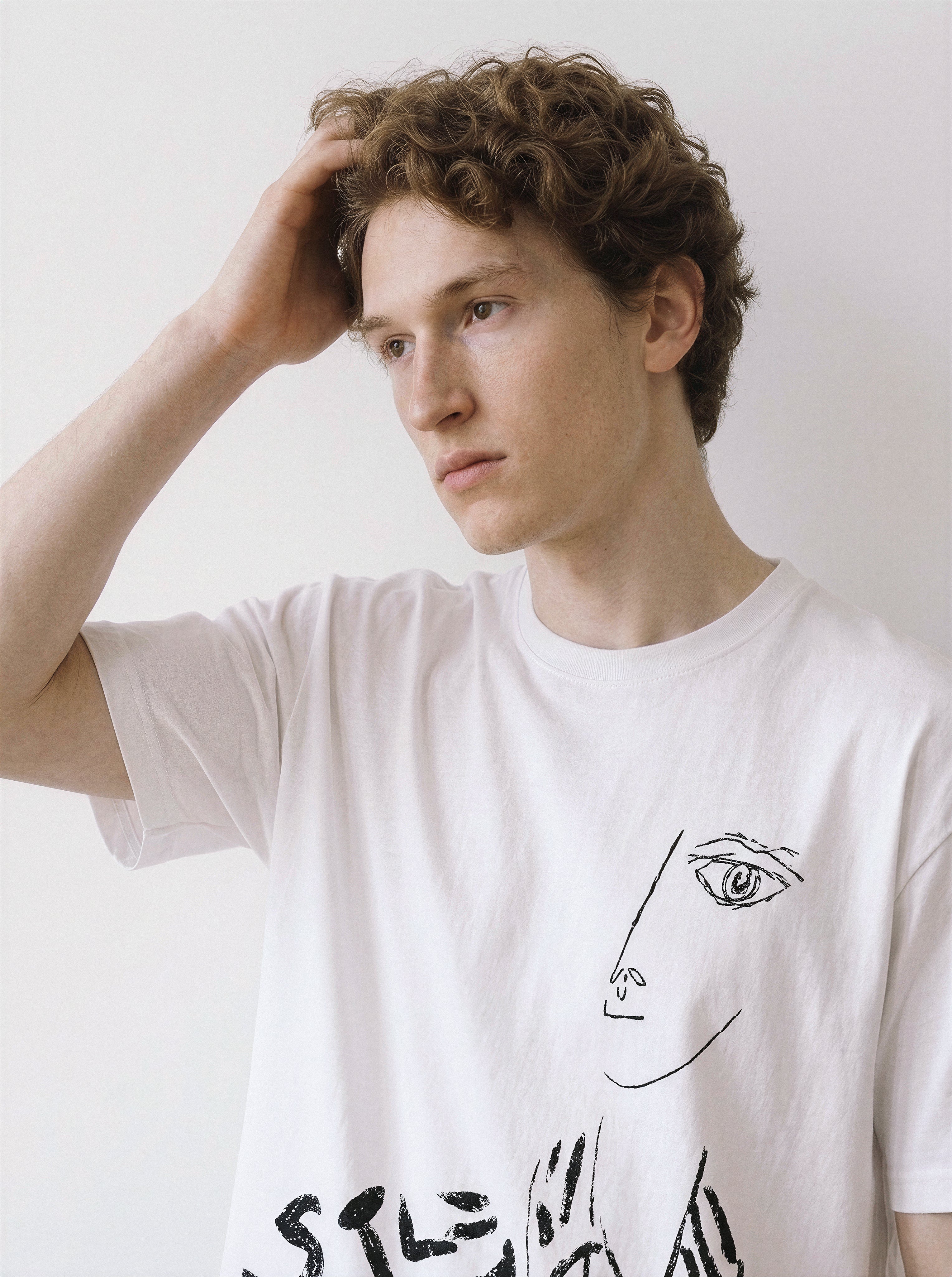 A male model with his hand in his hair posing in a white abstract one-eye face line art graphic tee, capturing a casual streetwear vibe with distinctive ink graphics.