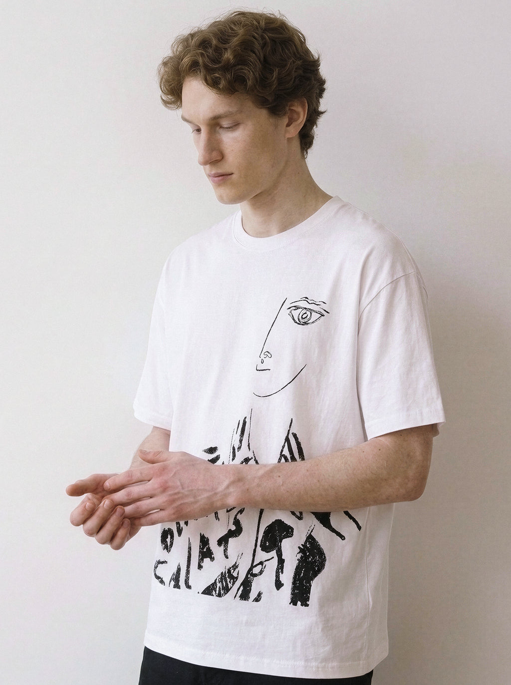 A male model looking down wearing a white abstract one-eye face line art graphic tee, showcasing a high-contrast independent manga style and minimalist streetwear look.