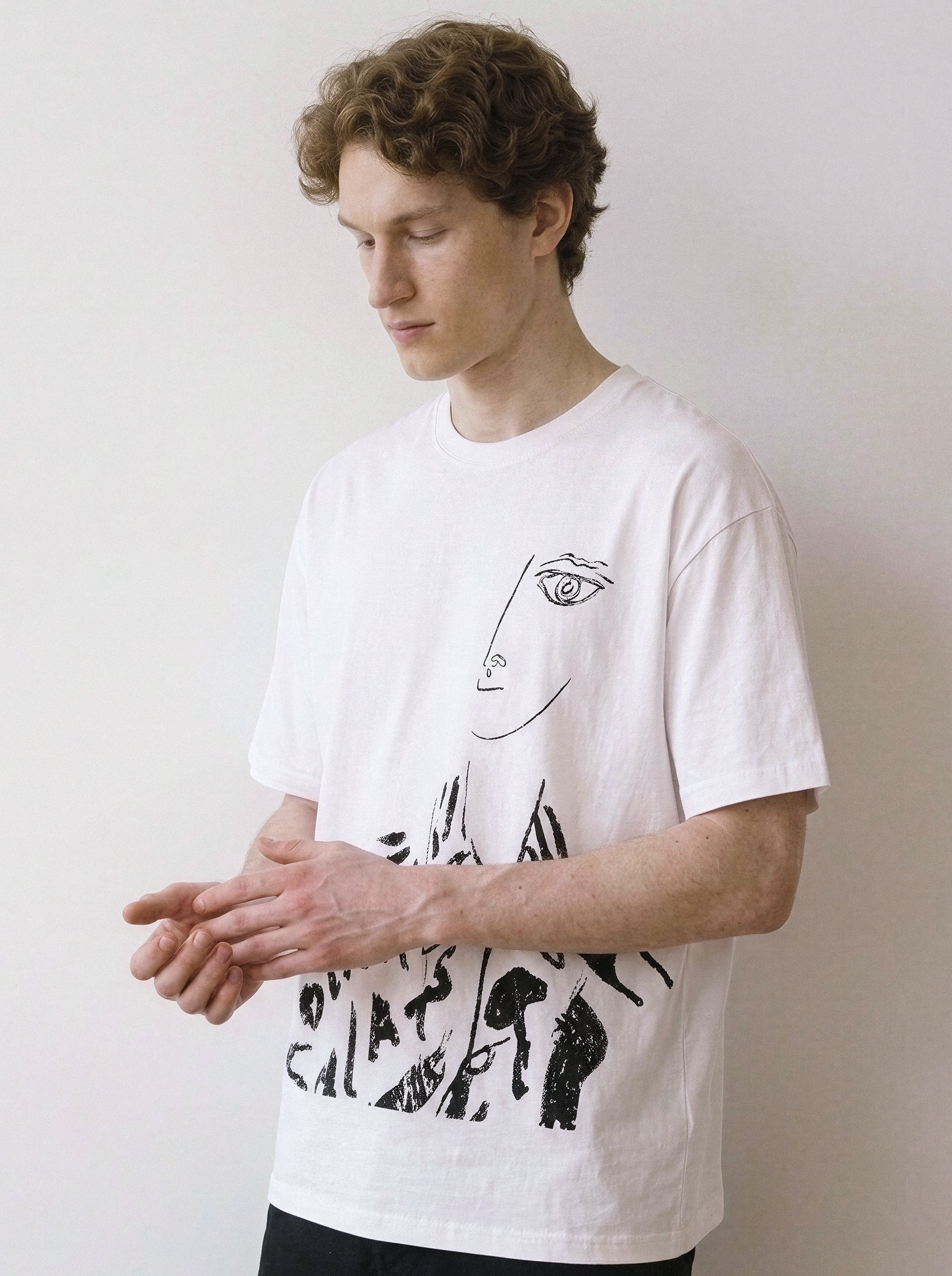 A male model looking down wearing a white abstract one-eye face line art graphic tee, showcasing a high-contrast independent manga style and minimalist streetwear look.