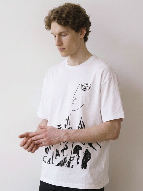 A male model looking down wearing a white abstract one-eye face line art graphic tee, showcasing a high-contrast independent manga style and minimalist streetwear look.