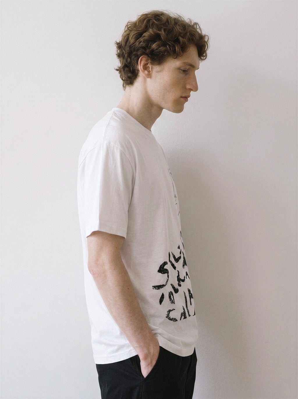 A side profile of a male model wearing a white abstract one-eye face line art graphic tee, emphasizing the relaxed unisex fit and clean independent manga aesthetic.