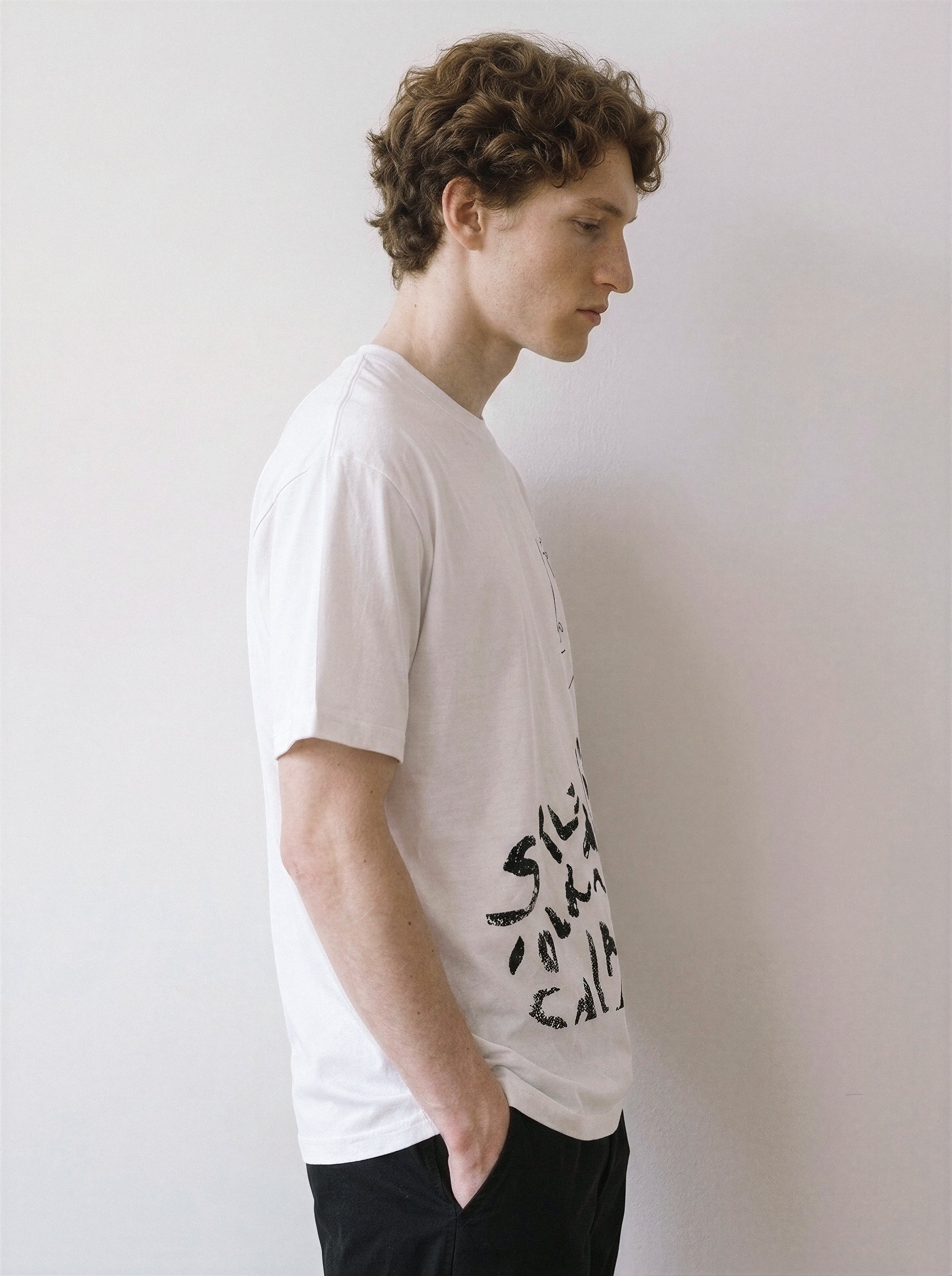 A side profile of a male model wearing a white abstract one-eye face line art graphic tee, emphasizing the relaxed unisex fit and clean independent manga aesthetic.