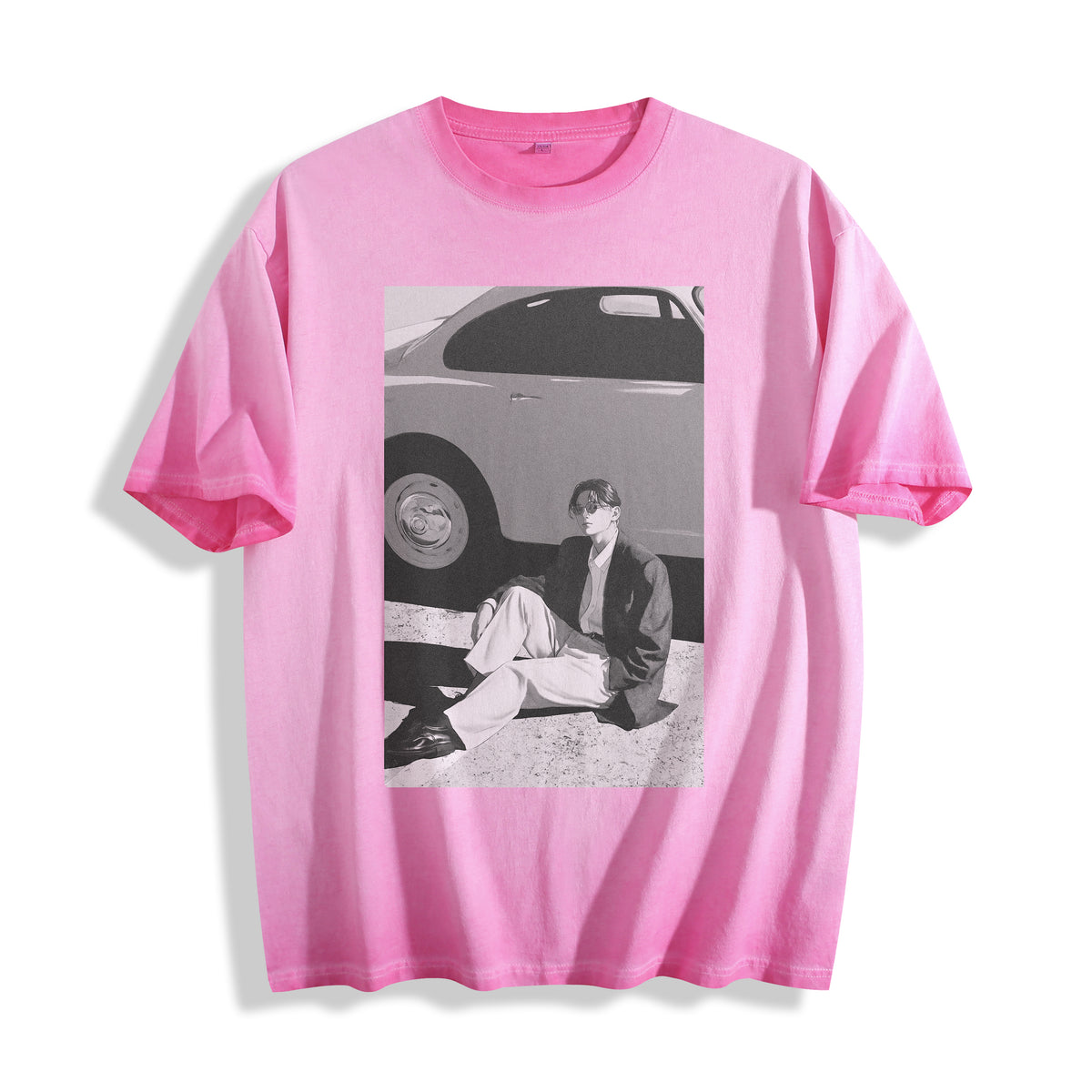 A pink short-sleeve crewneck t-shirt featuring a rectangular 90s retro black and white photography print on the front showing a person sitting next to a car, displayed against a clean white background.