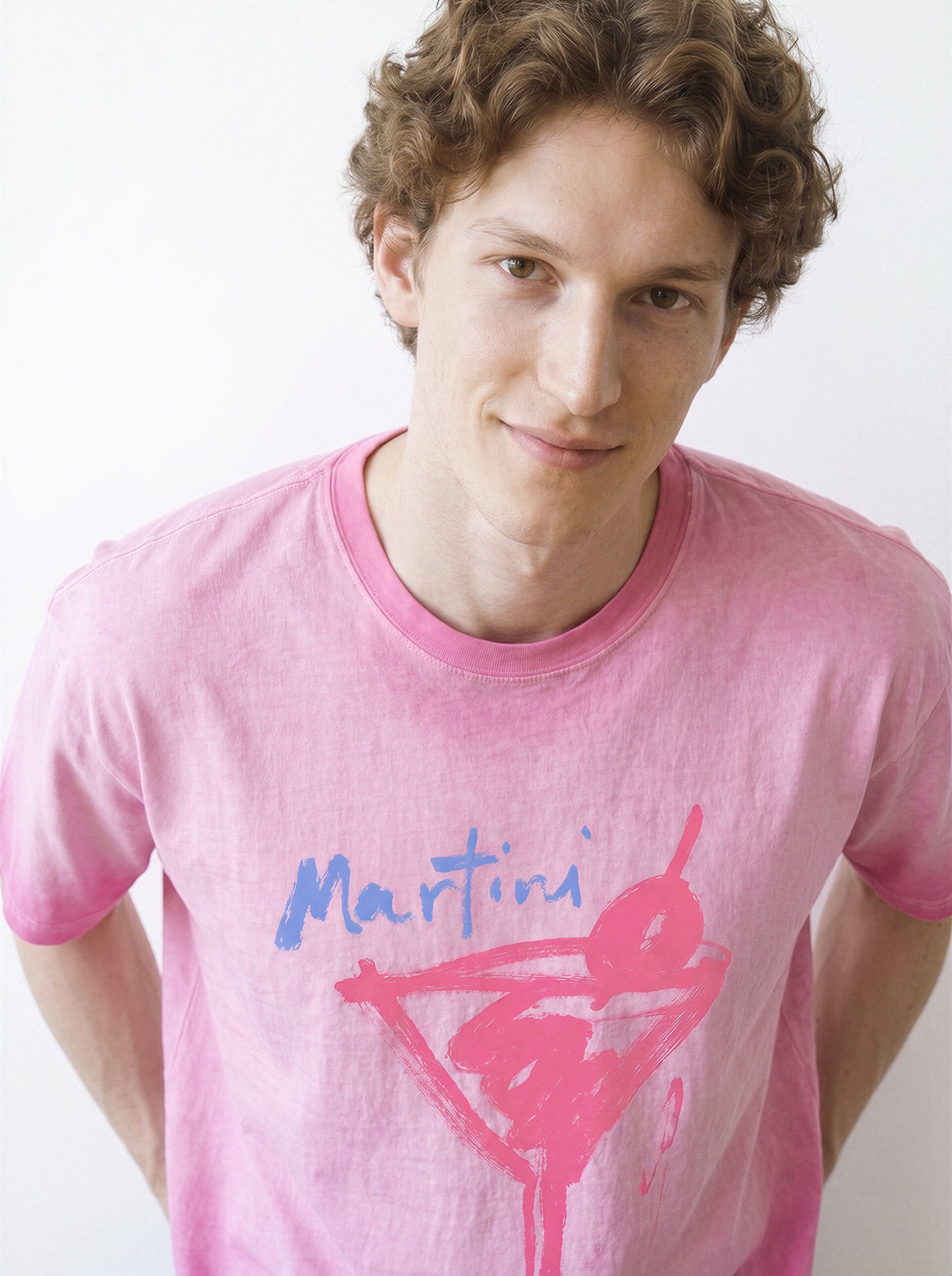 Front view of a male model smiling at the camera, demonstrating the relaxed drape of an aura pink structured streetwear t-shirt with a raw ink martini glass design.