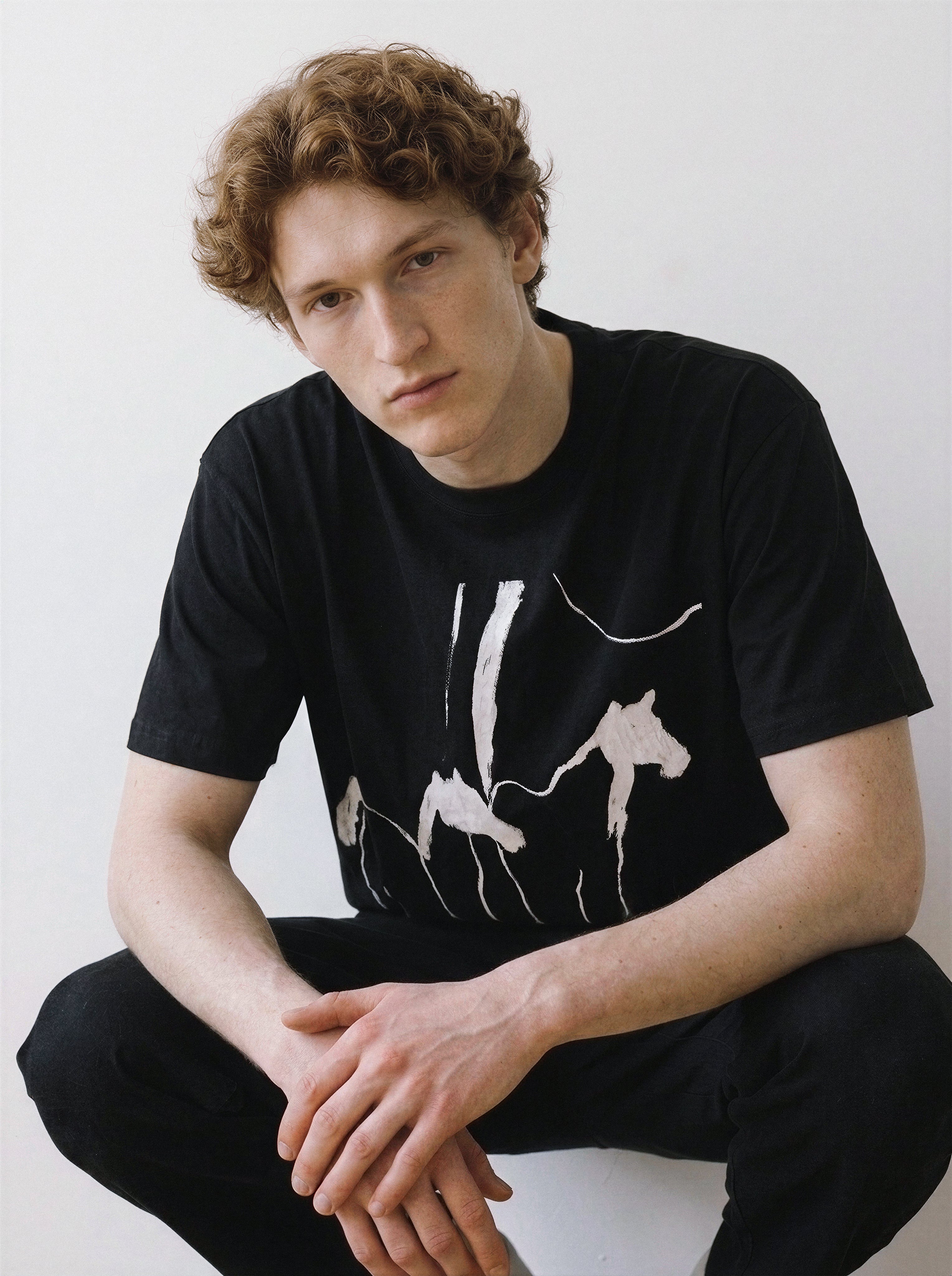 Front portrait of a male model crouching with clasped hands, wearing a black oversized graphic tee featuring a raw abstract brush wild horses print.