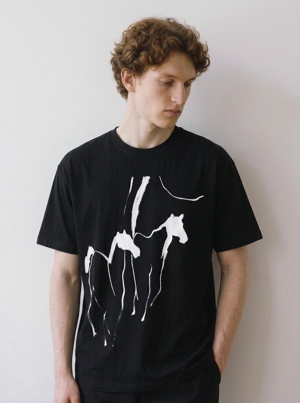 A male model looking down, demonstrating the relaxed drape of a black oversized streetwear t-shirt with a bold sweeping brush stroke horses design.