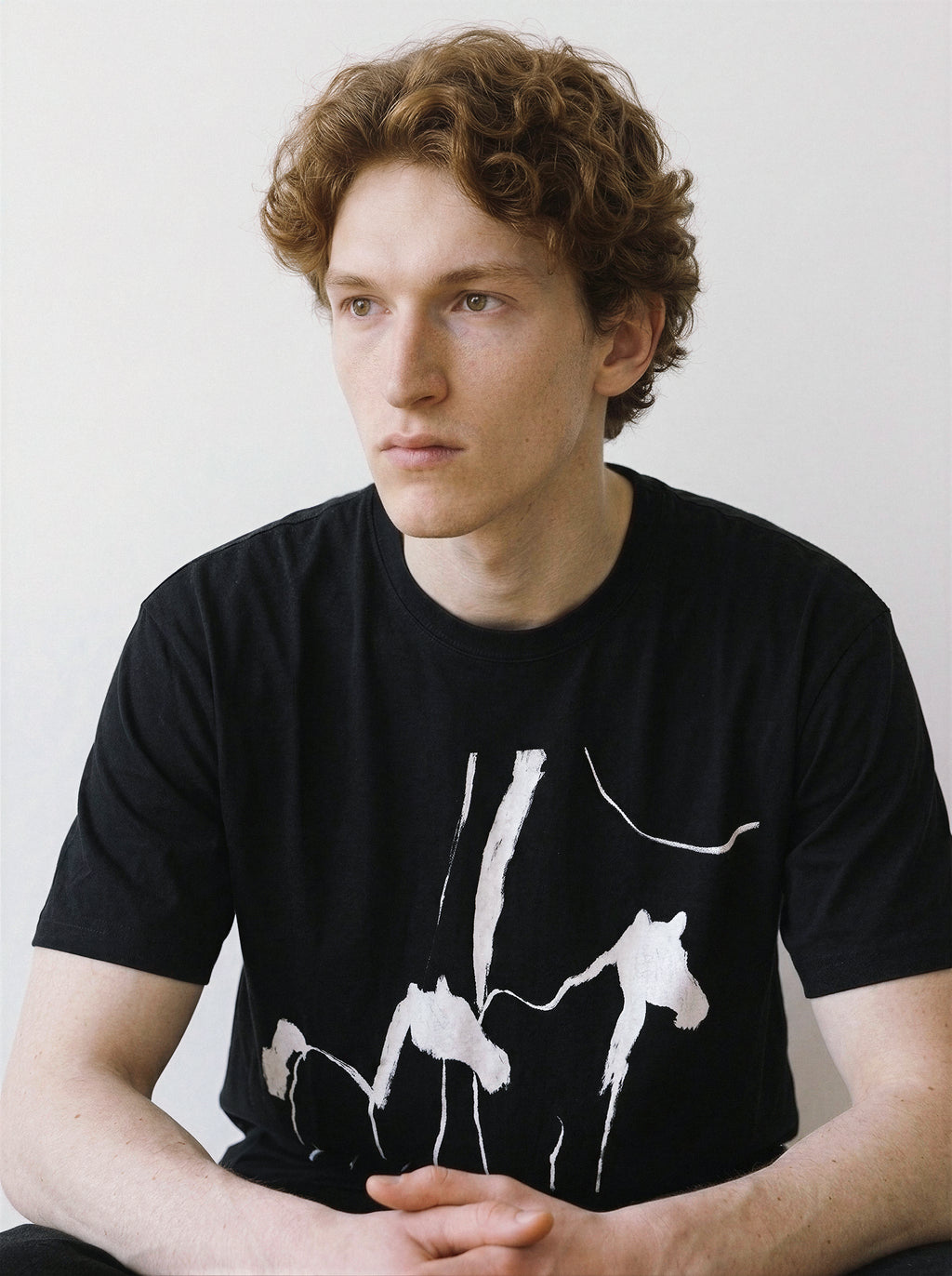 Front view of a model sitting and leaning slightly forward, highlighting the 180g premium cotton fit of a black tee with an expressive abstract wild horses illustration.