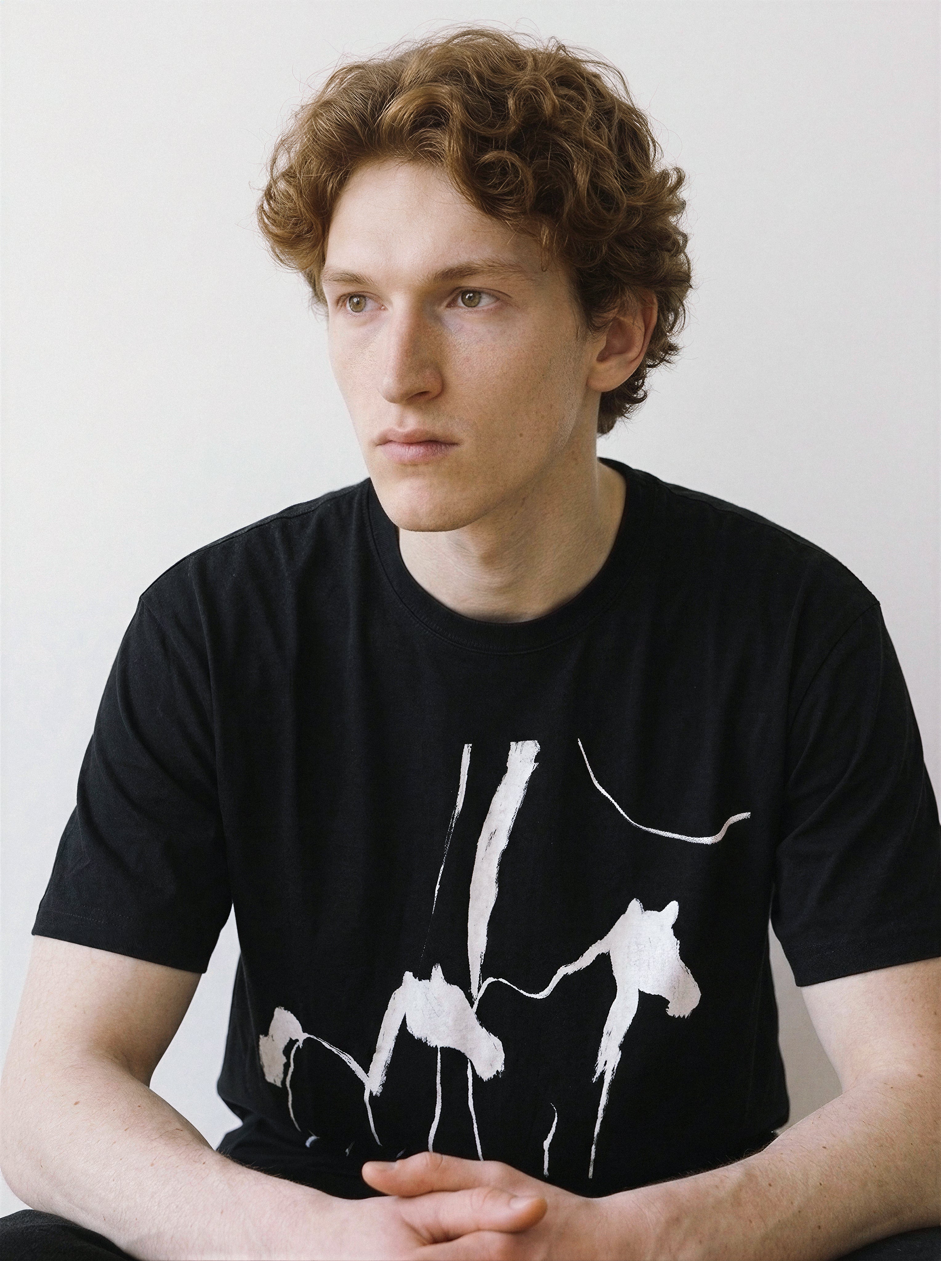 Front view of a model sitting and leaning slightly forward, highlighting the 180g premium cotton fit of a black tee with an expressive abstract wild horses illustration.