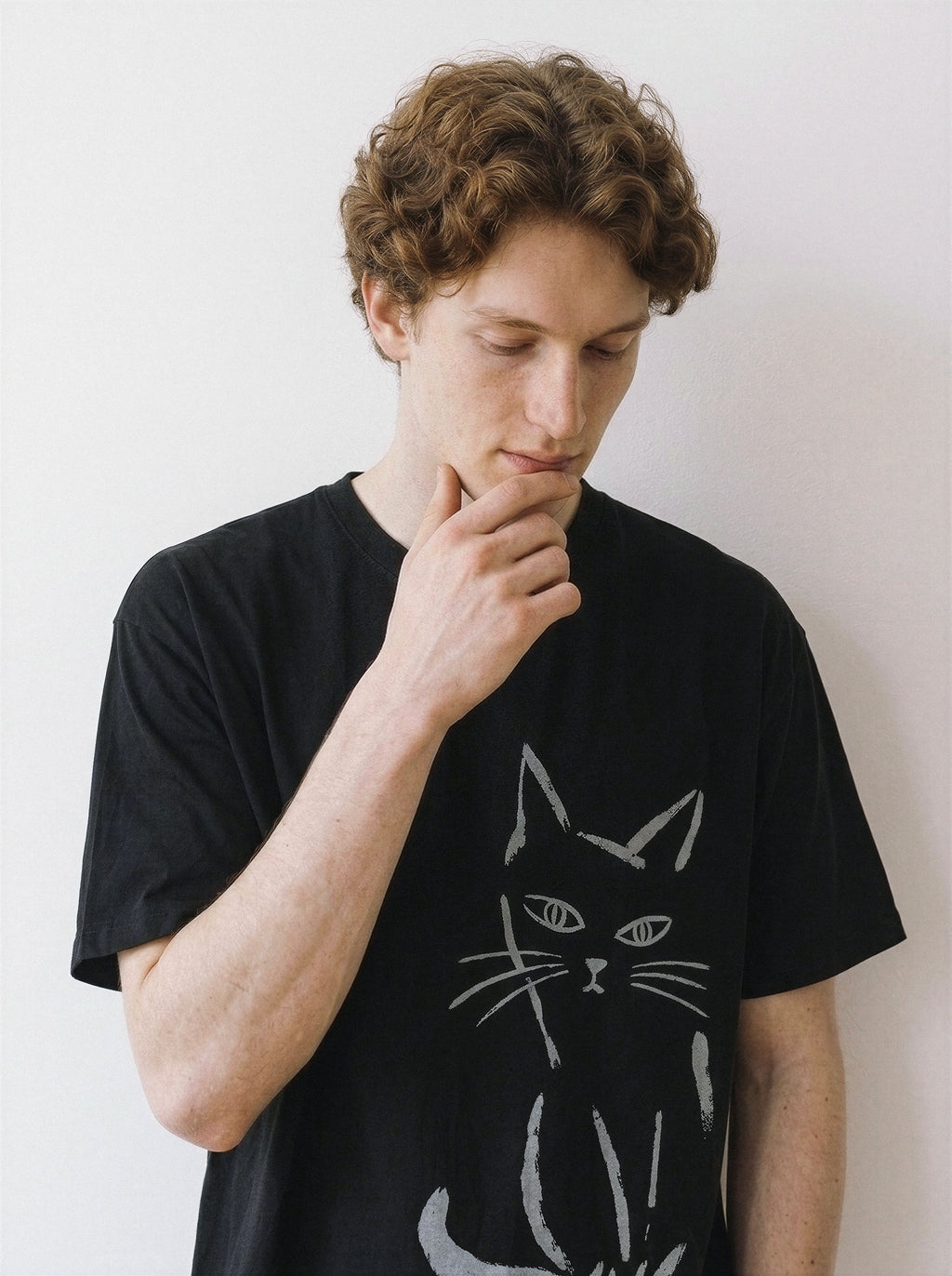 Front portrait of a male model looking down and resting his hand on his chin, wearing a black oversized graphic tee featuring a minimalist paint stroke judgmental cat.
