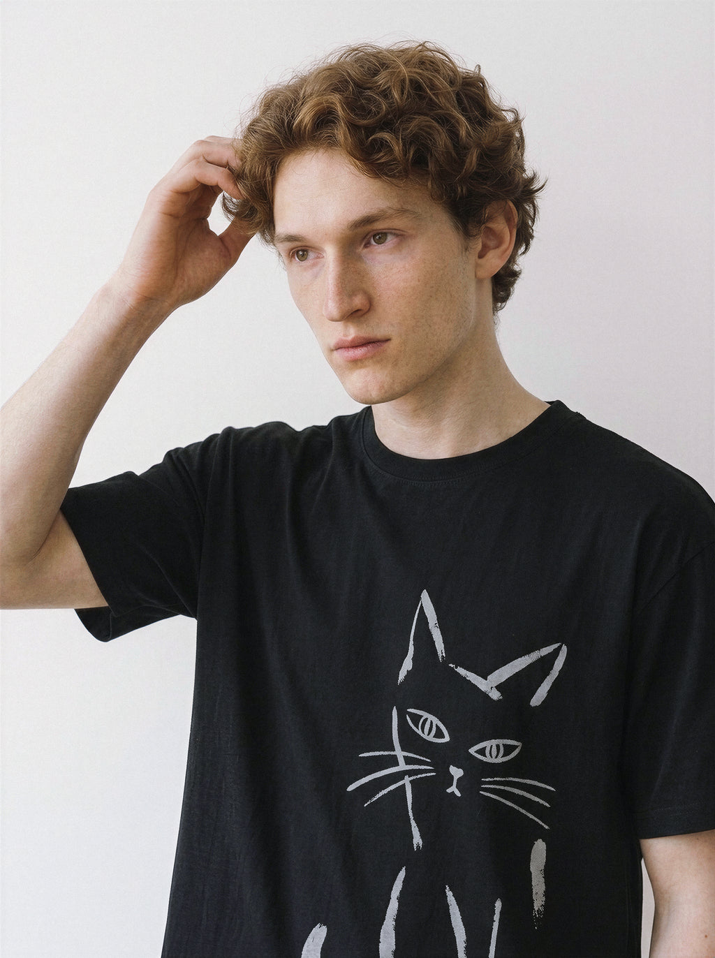 Front view of a male model with his hand in his hair, wearing a black 180g premium cotton tee highlighting a messy paint stroke feline design.