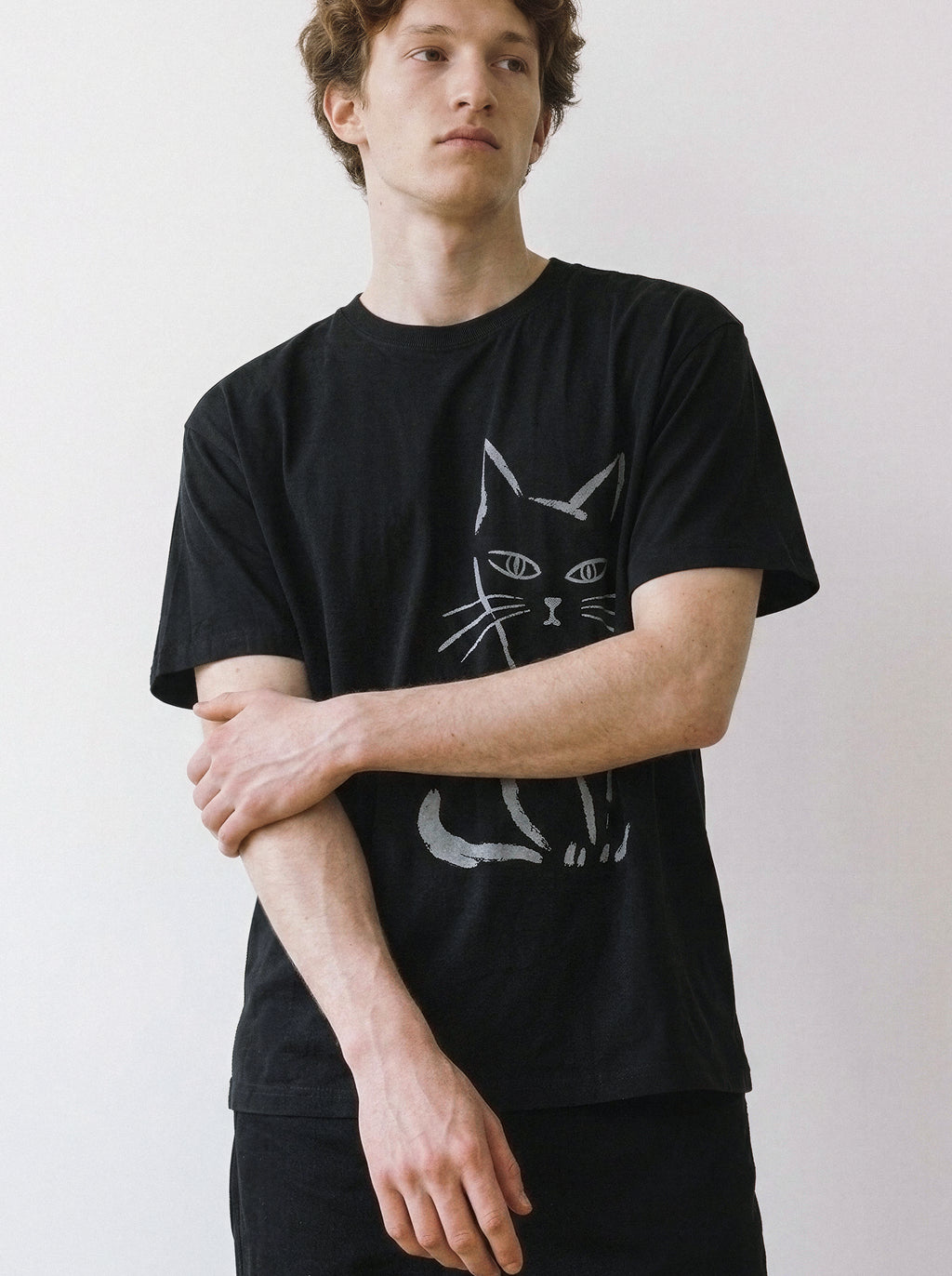 A male model looking away while holding his arm, highlighting the relaxed drape of a black oversized streetwear t-shirt with a raw side-eye cat line art print.
