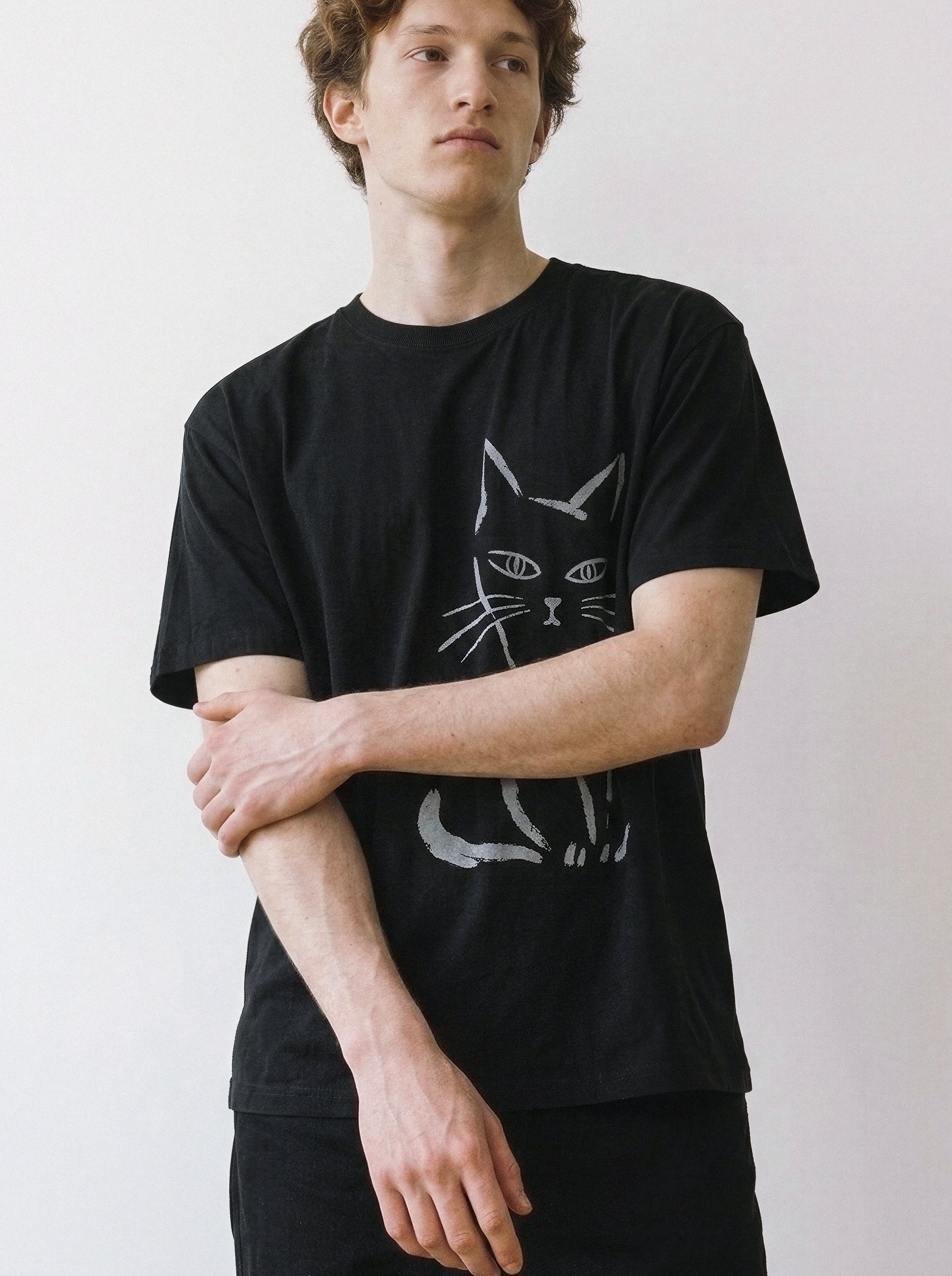 A male model looking away while holding his arm, highlighting the relaxed drape of a black oversized streetwear t-shirt with a raw side-eye cat line art print.
