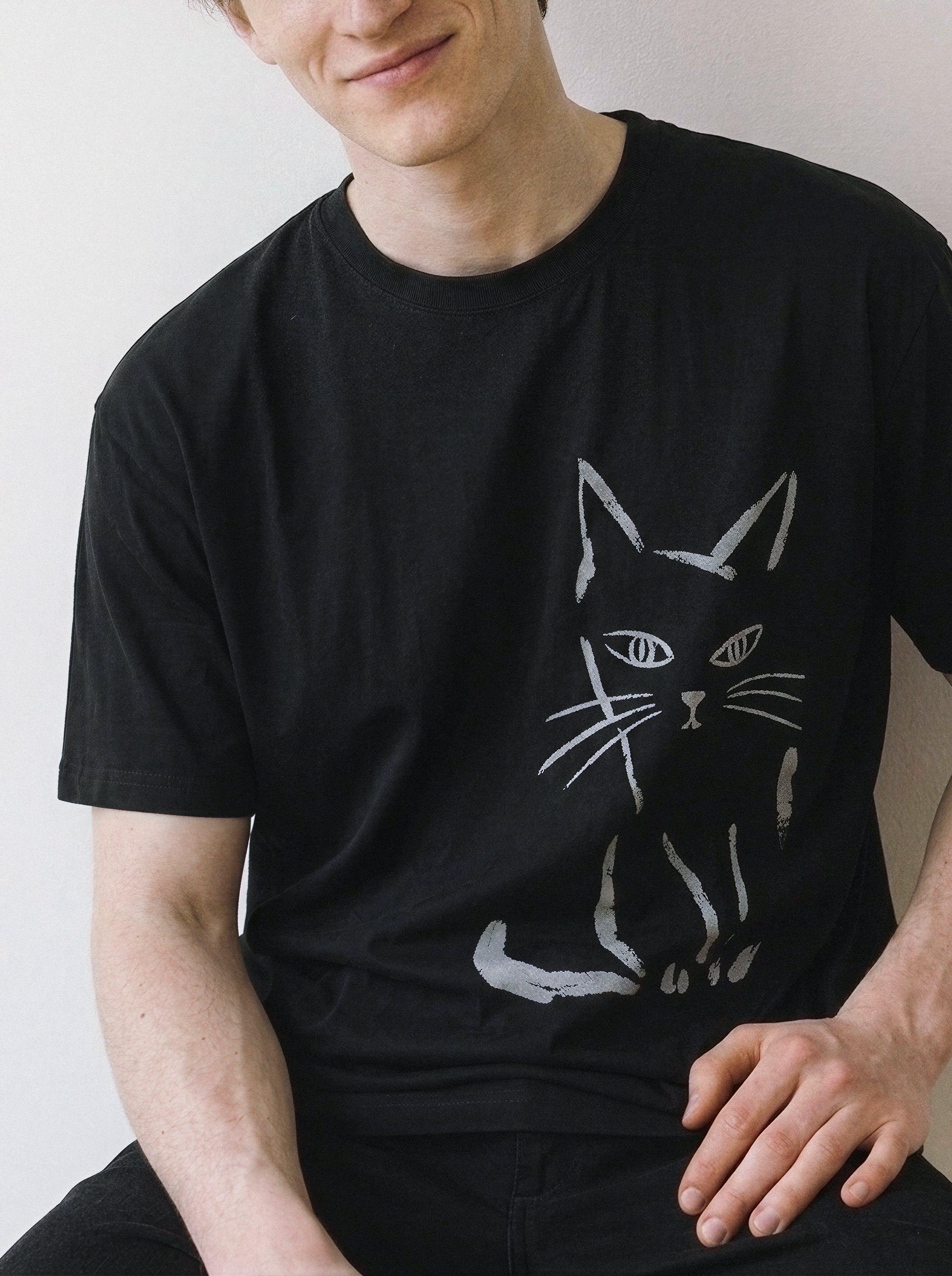 A male model sitting and looking directly at the camera, showcasing a black oversized graphic tee with a bold judgmental cat illustration.