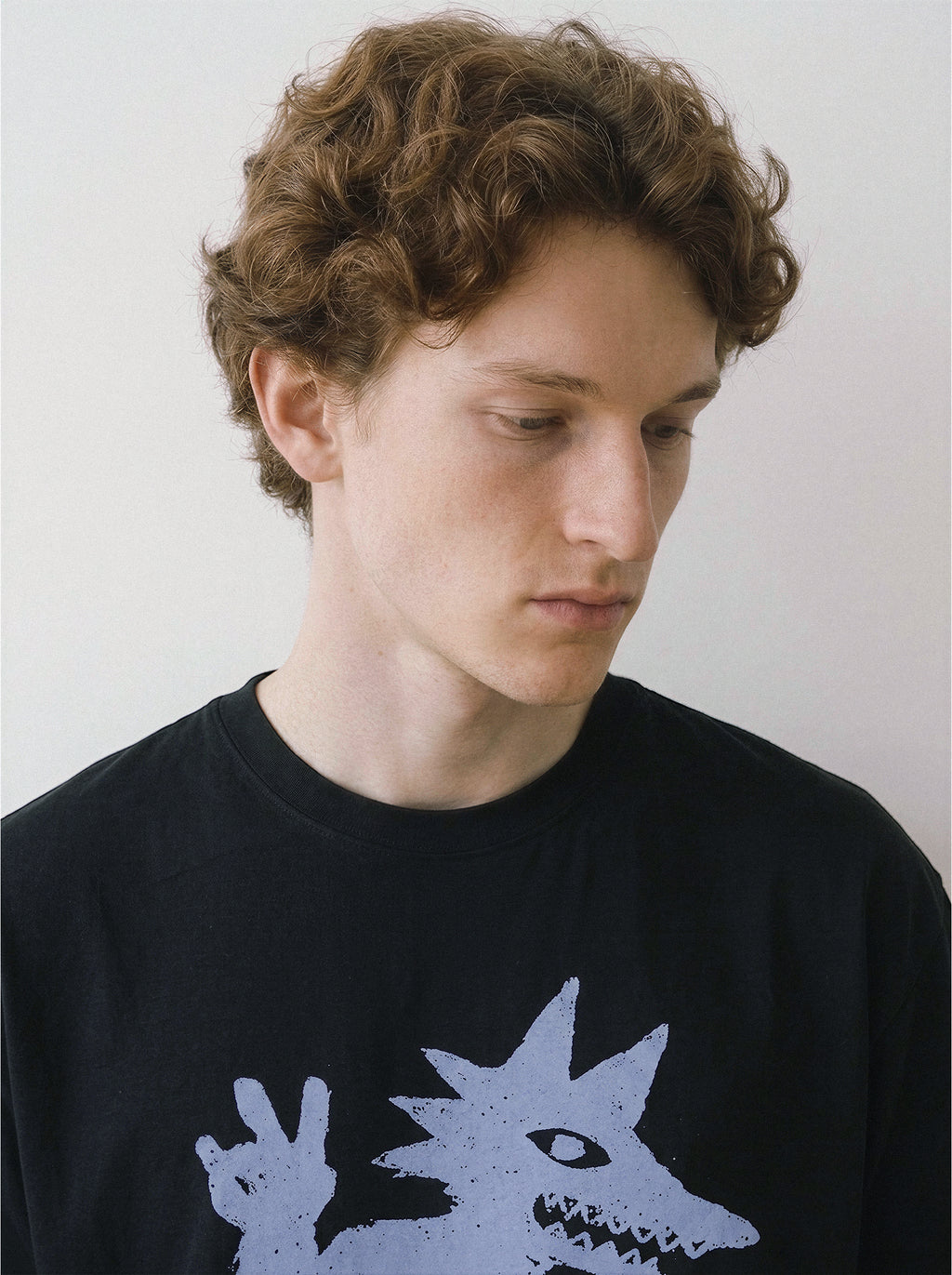Close-up chest view of a male model looking down, emphasizing the sneaker-wearing monster throwing a peace sign on a black oversized tee. 