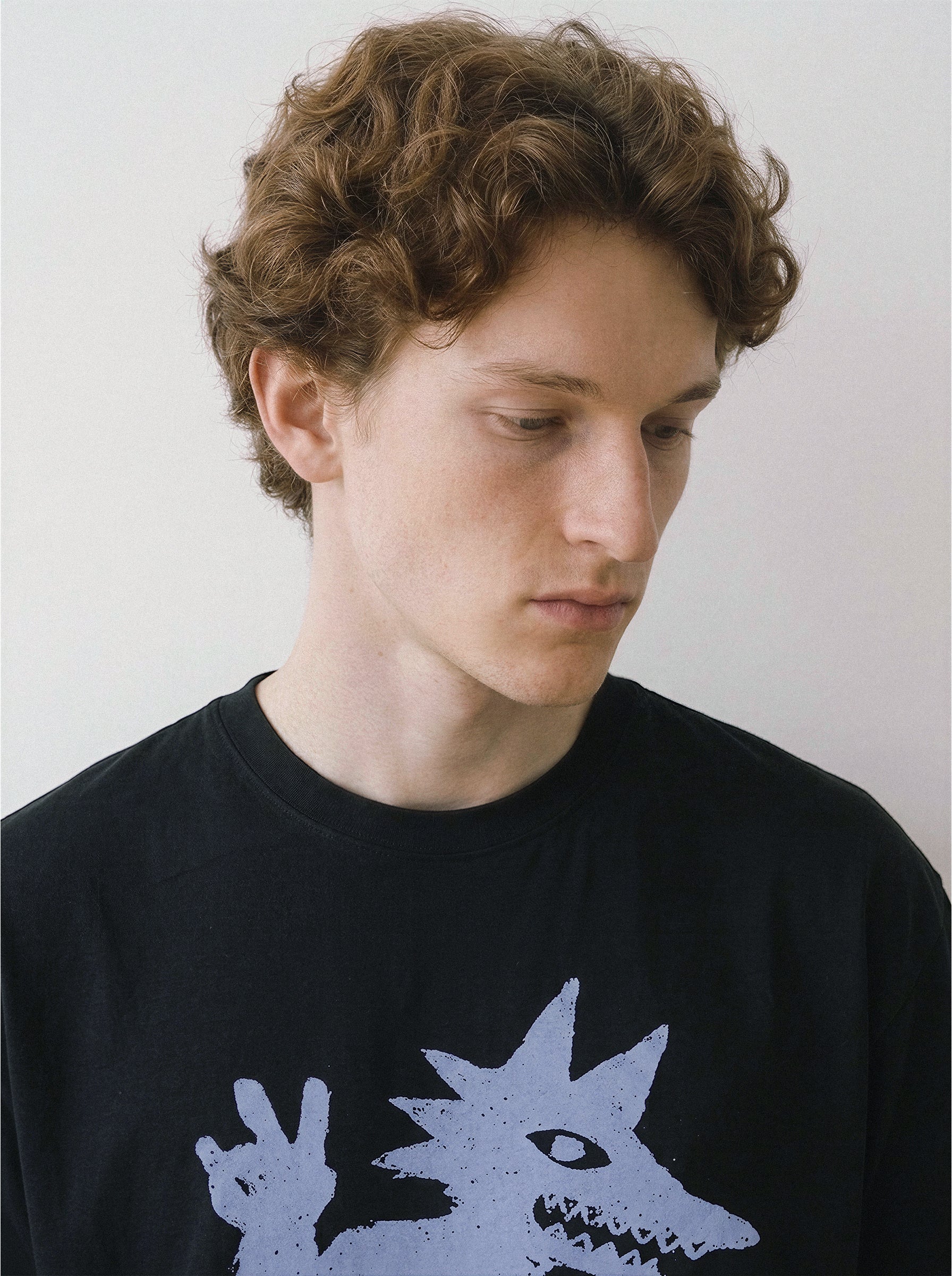 Close-up chest view of a male model looking down, emphasizing the sneaker-wearing monster throwing a peace sign on a black oversized tee. 
