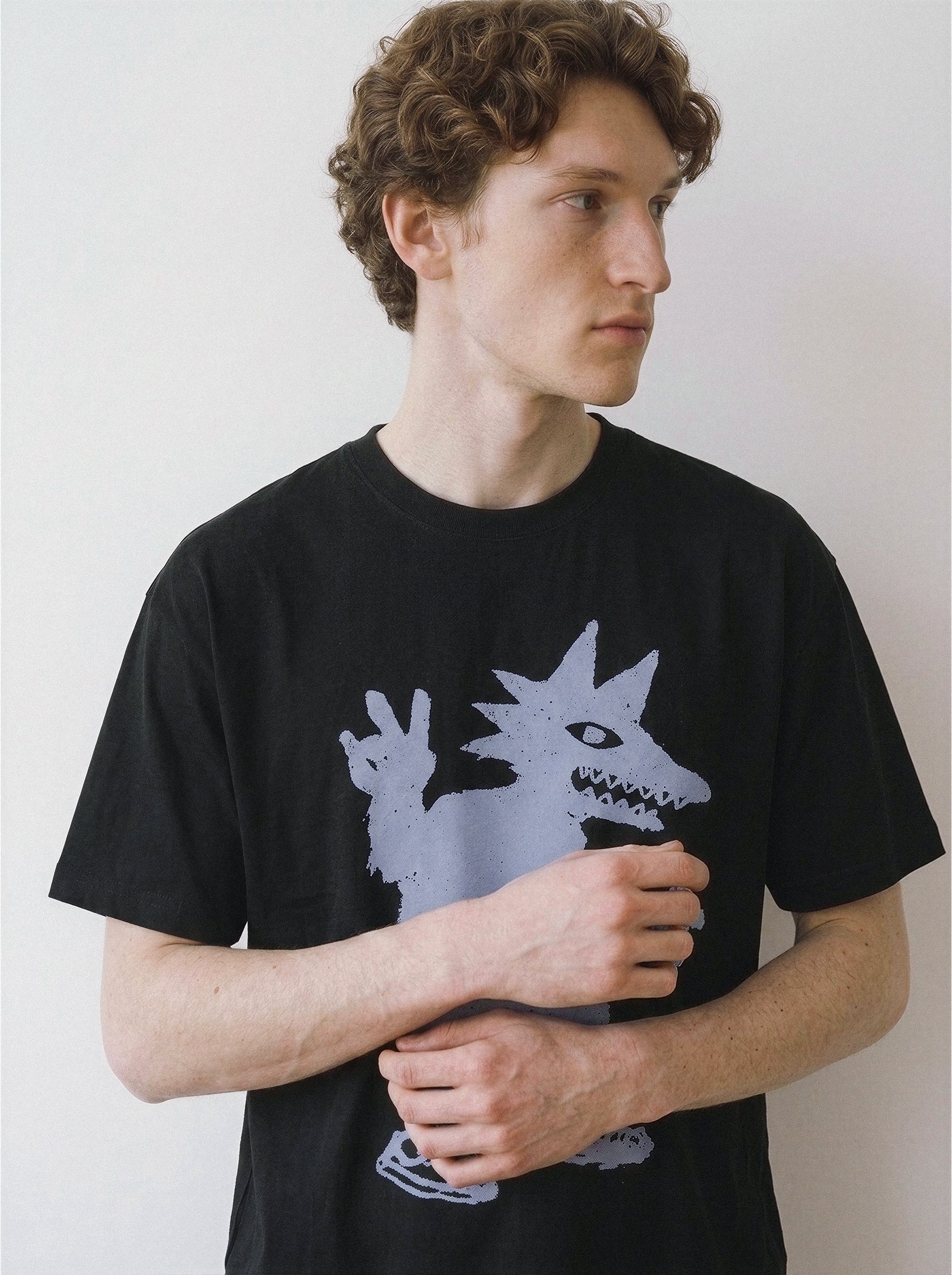 Front portrait of a male model looking away with hands loosely clasped, wearing a black oversized graphic tee featuring a skate-culture peace sign monster doodle.