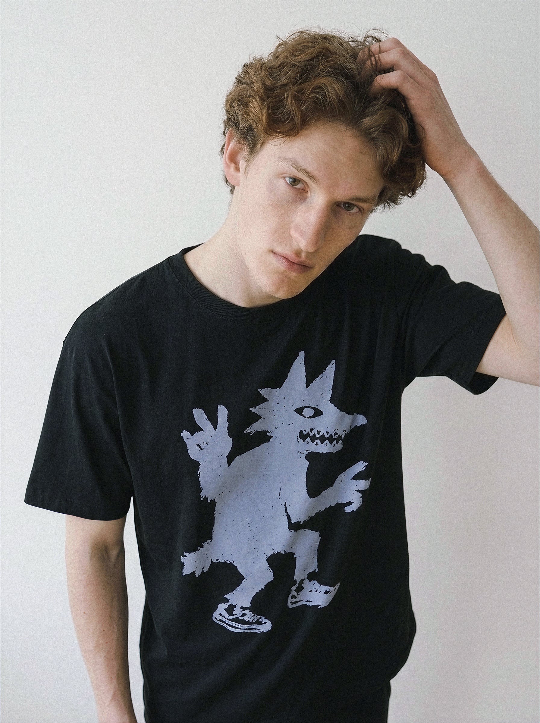 A young man with curly light brown hair wearing a black t-shirt with a light gray graphic of a spiky-haired cartoon character, posing against a plain off-white wall.