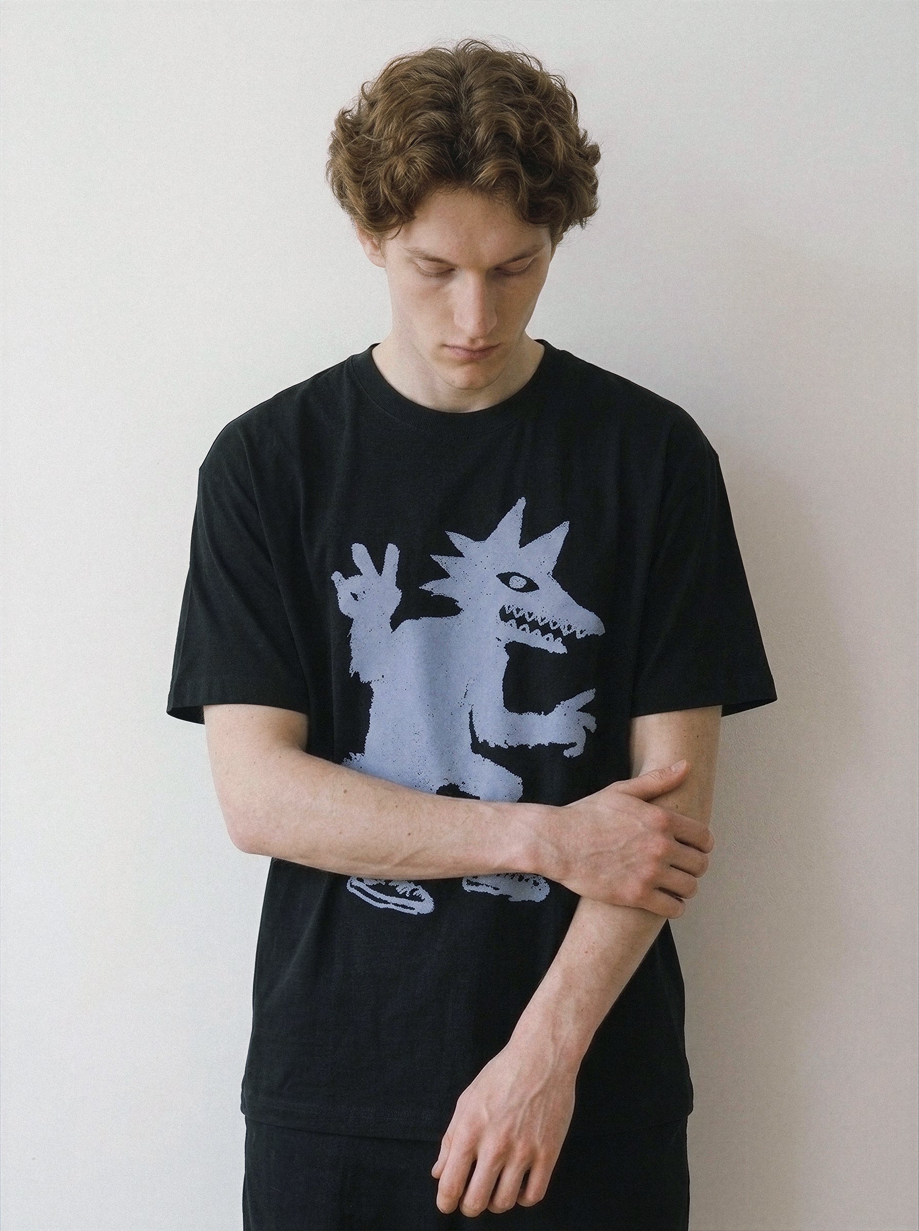 A male model looking down with hands resting in front, highlighting the relaxed drape of a black oversized graphic tee with a peace sign creature.