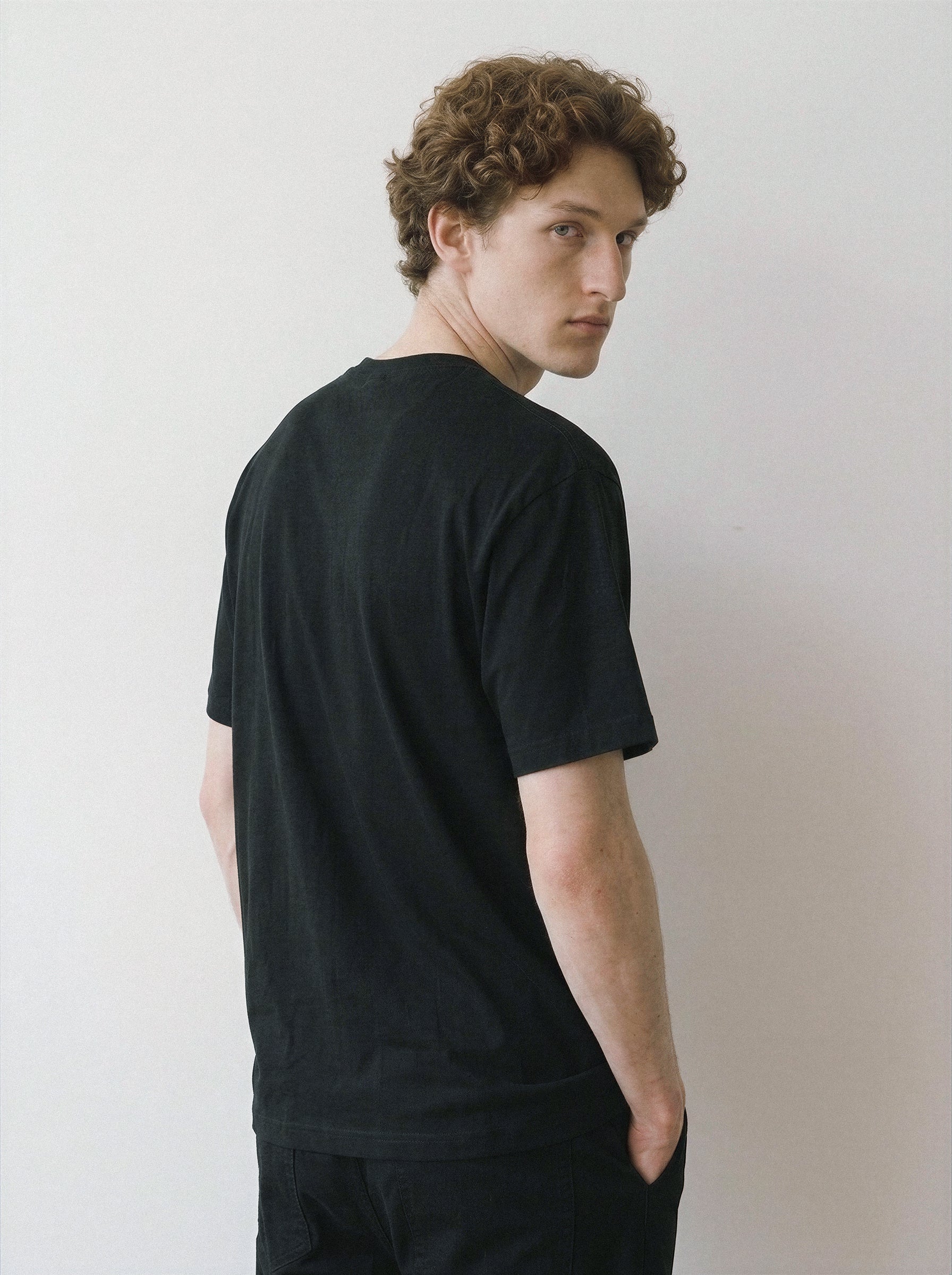 A man with curly brown hair wearing a black short-sleeve crewneck t-shirt, photographed from the back and side against a plain light gray wall.