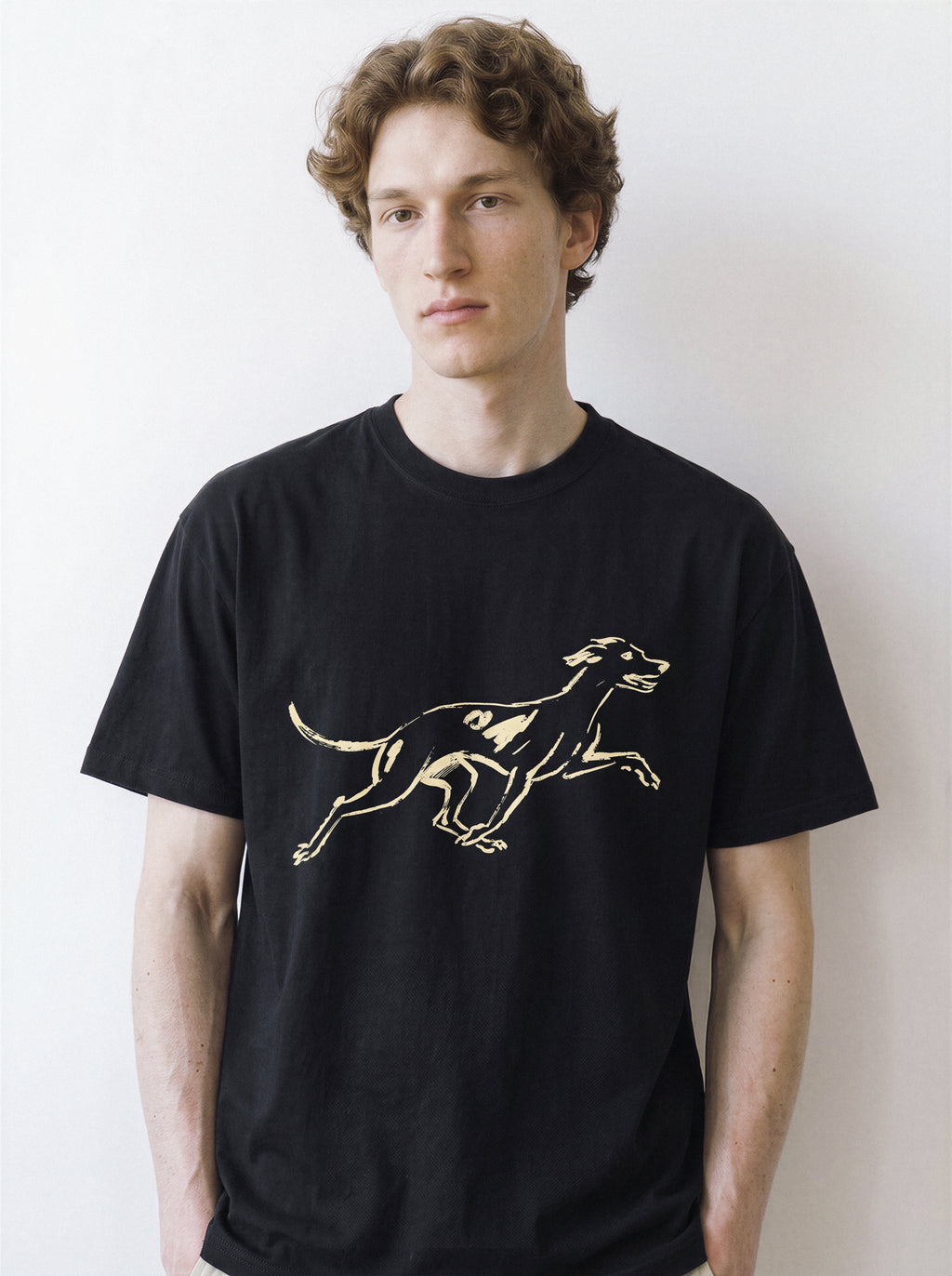 Front portrait of a male model looking directly at the camera, wearing a black oversized graphic tee featuring a pure line art print of a running dog with a surprised expression.