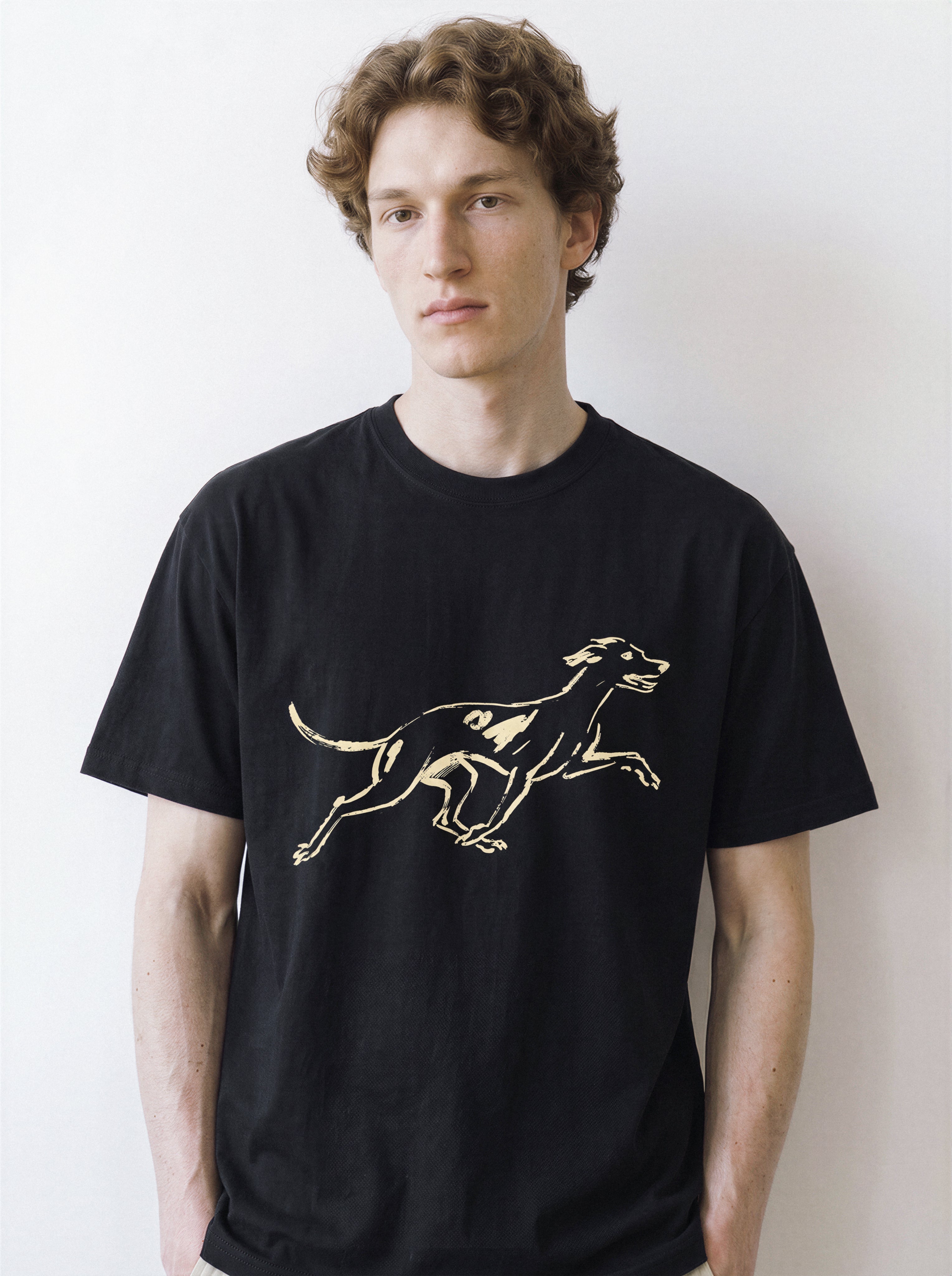 Front portrait of a male model looking directly at the camera, wearing a black oversized graphic tee featuring a pure line art print of a running dog with a surprised expression.