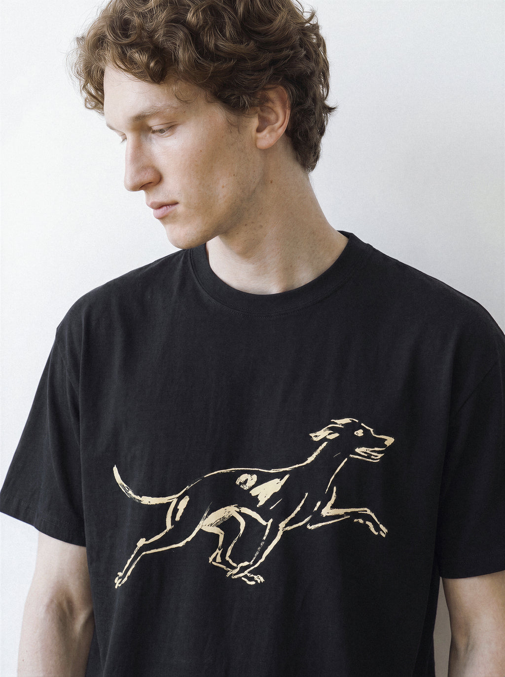 A male model looking down, demonstrating the relaxed drape of a black 180g oversized streetwear t-shirt with a minimalist surprised hound brushstroke design.