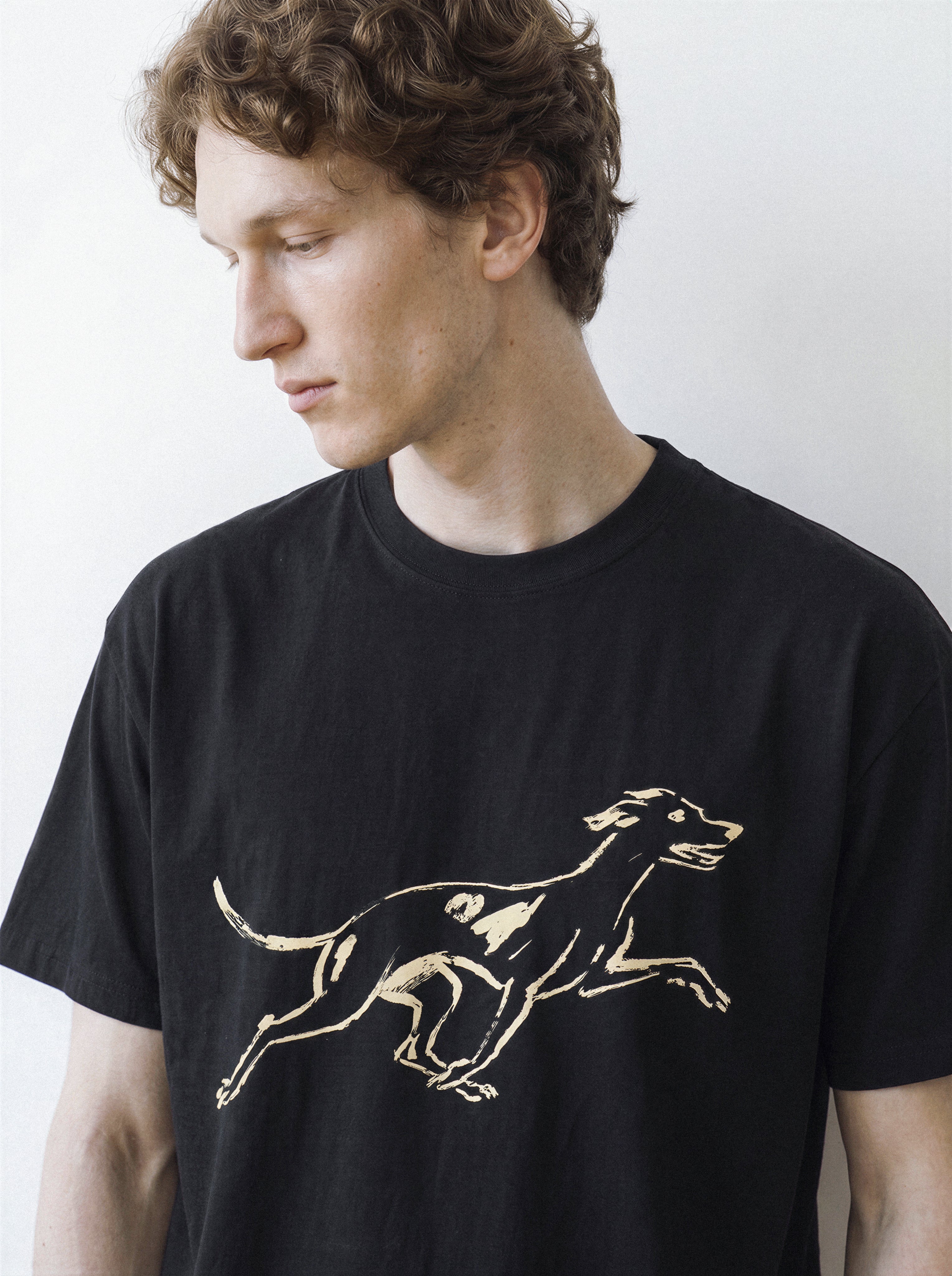 A male model looking down, demonstrating the relaxed drape of a black 180g oversized streetwear t-shirt with a minimalist surprised hound brushstroke design.