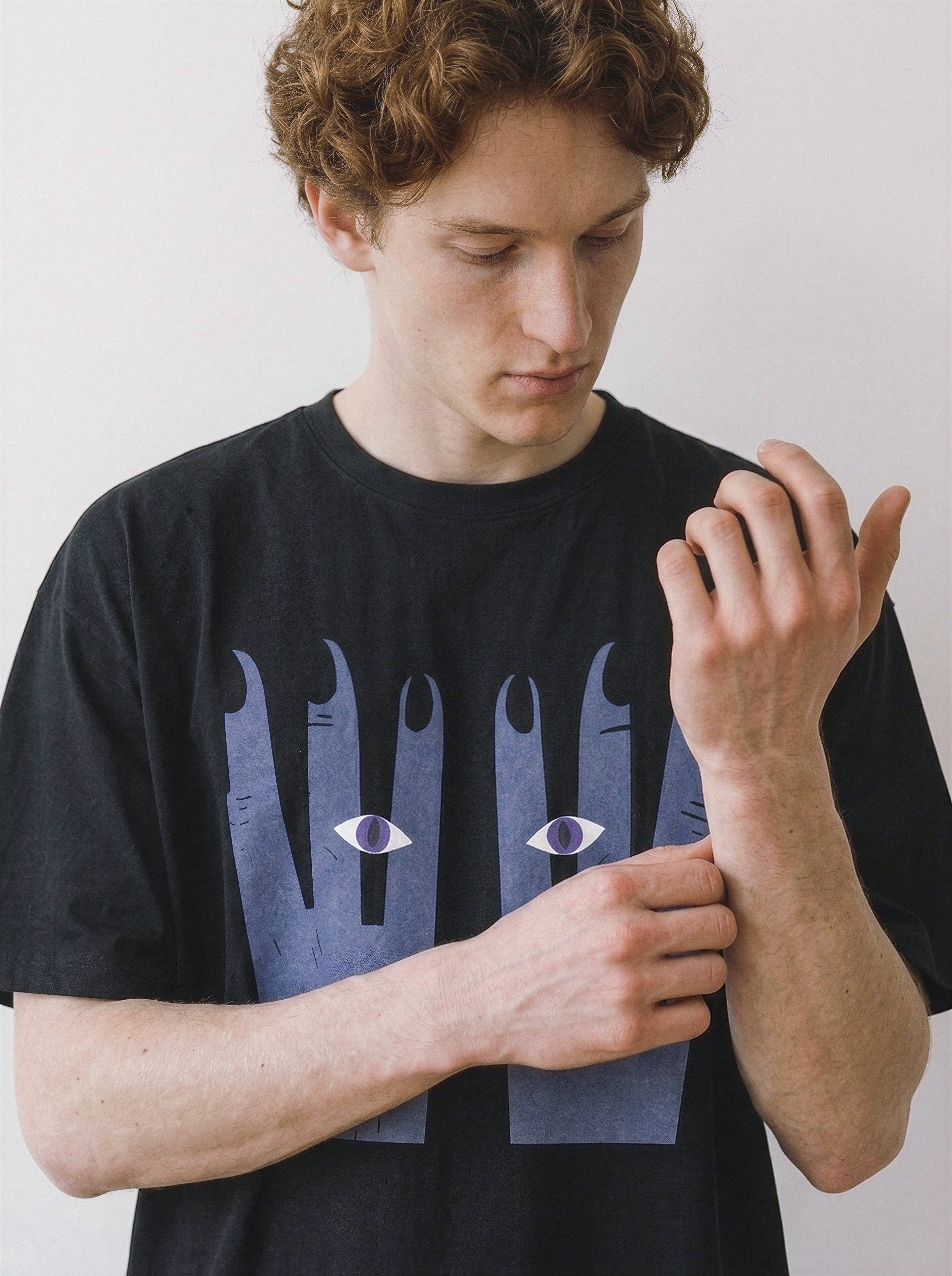 A male model adjusting his sleeve, wearing a black 180g oversized cotton tee highlighting a surreal hands and eyes graphic design.