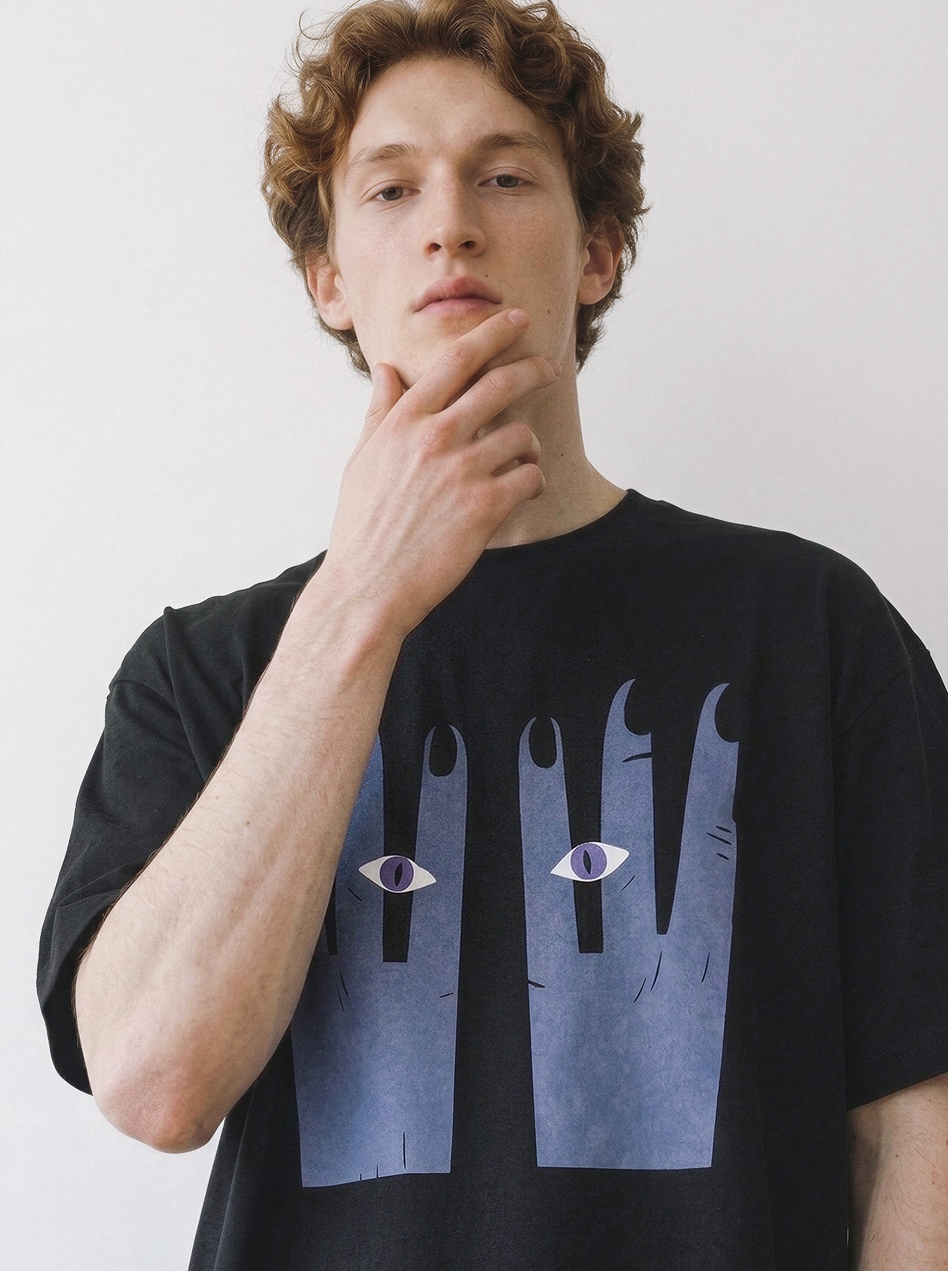 Front portrait of a male model resting his hand on his chin, wearing a black oversized graphic tee featuring a bold surreal hands and eyes art print.