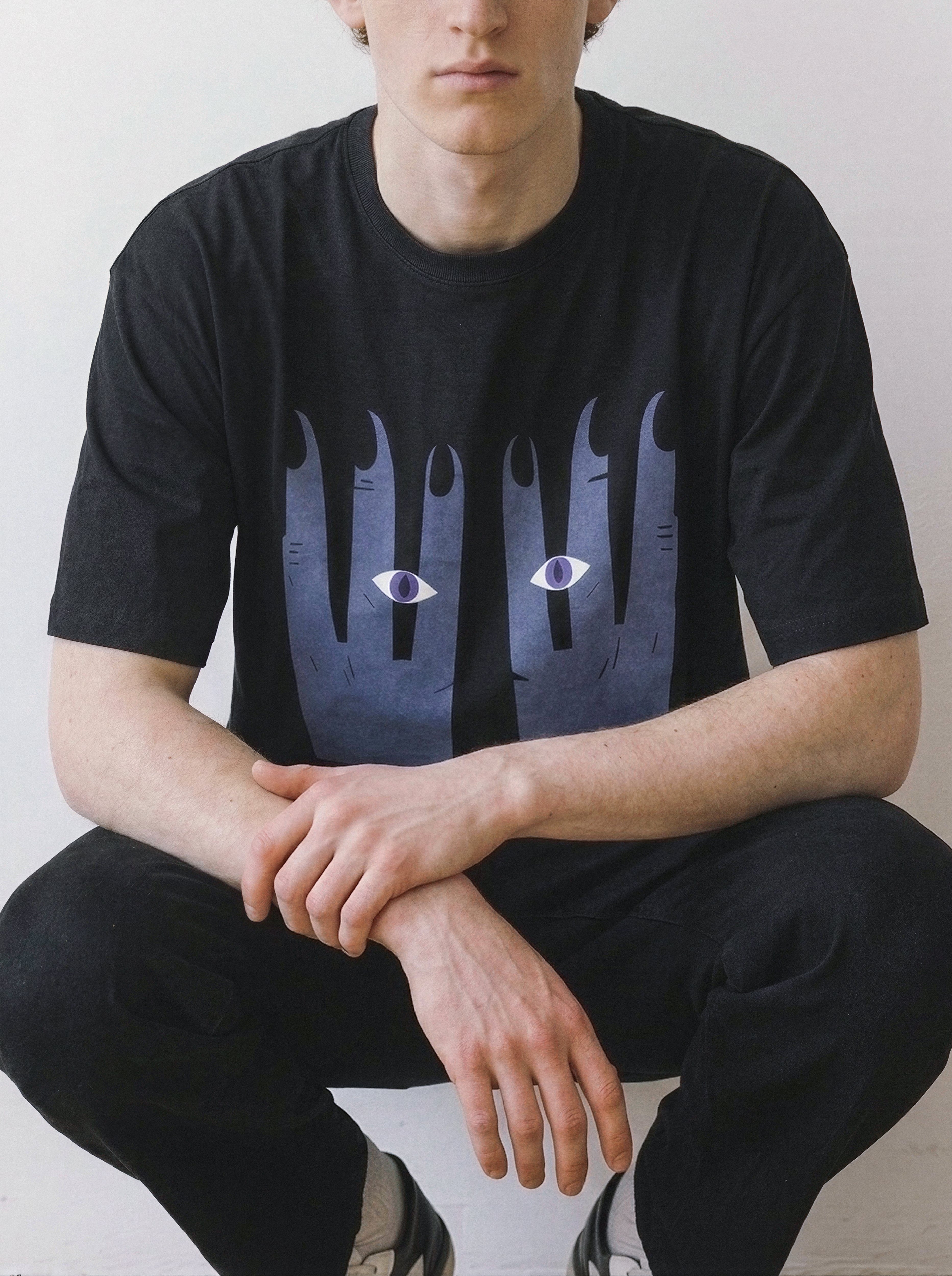 A person wearing a black short-sleeve graphic T-shirt with a large dark blue and gray abstract hand-like print on the front, featuring two small white-and-purple eyes. The person is seated against a plain light wall.