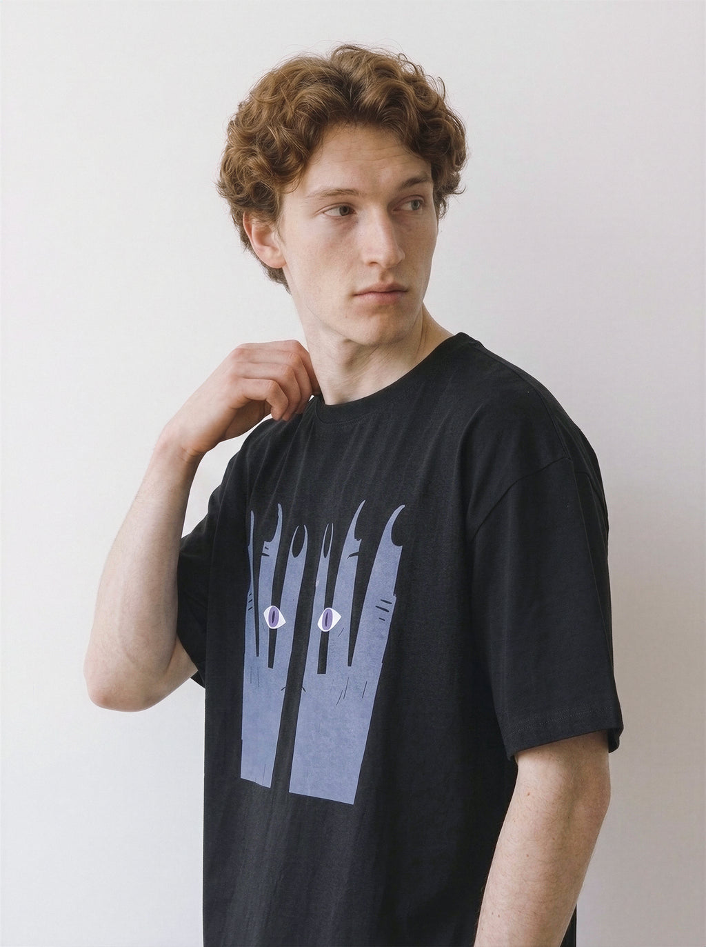 A male model looking away while touching his neck, showcasing the fit of a black oversized t-shirt with a unique surreal illustration.