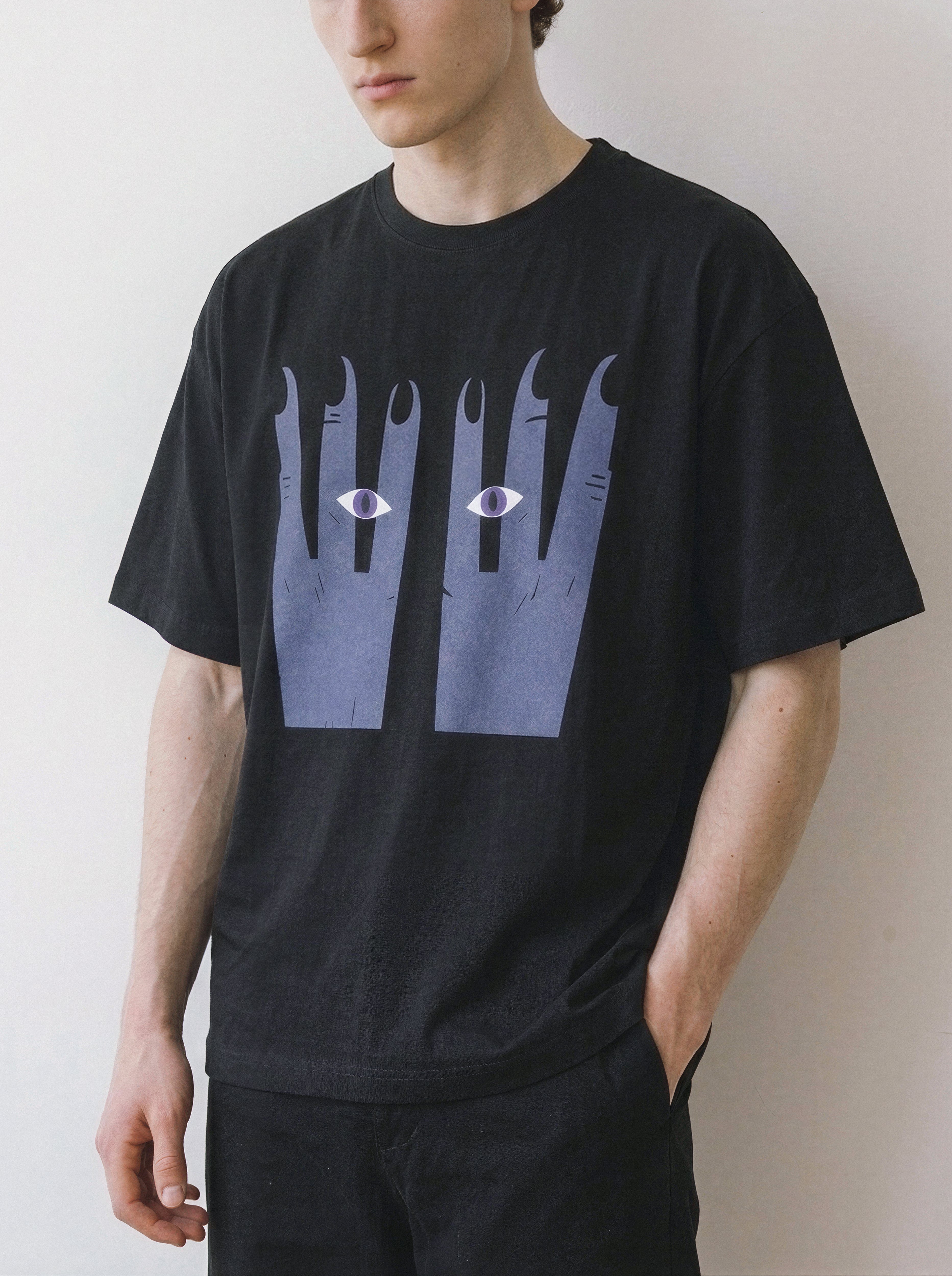 A man wearing a black crewneck t-shirt with a graphic print of two blue-grey hands with eyes on the front, posed against a plain off-white wall.
