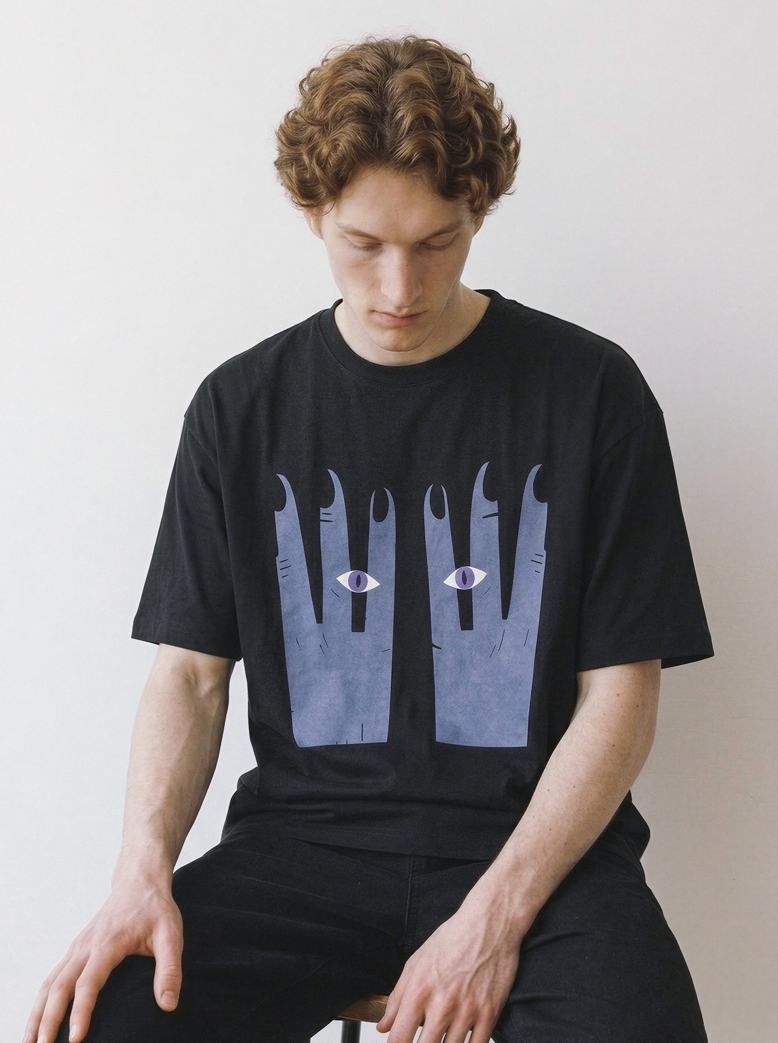 A male model looking down while sitting, showcasing a black oversized t-shirt with a striking surreal hands and eyes illustration.