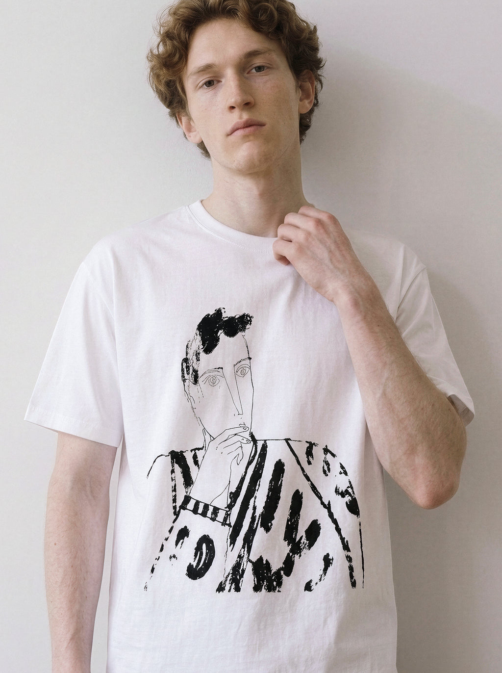 A male model leaning against a wall wearing a white blank stare portrait line art graphic tee, showcasing the heavy stroke sketch and relaxed unisex fit.