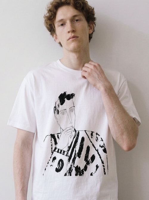 A male model leaning against a wall wearing a white blank stare portrait line art graphic tee, showcasing the heavy stroke sketch and relaxed unisex fit.