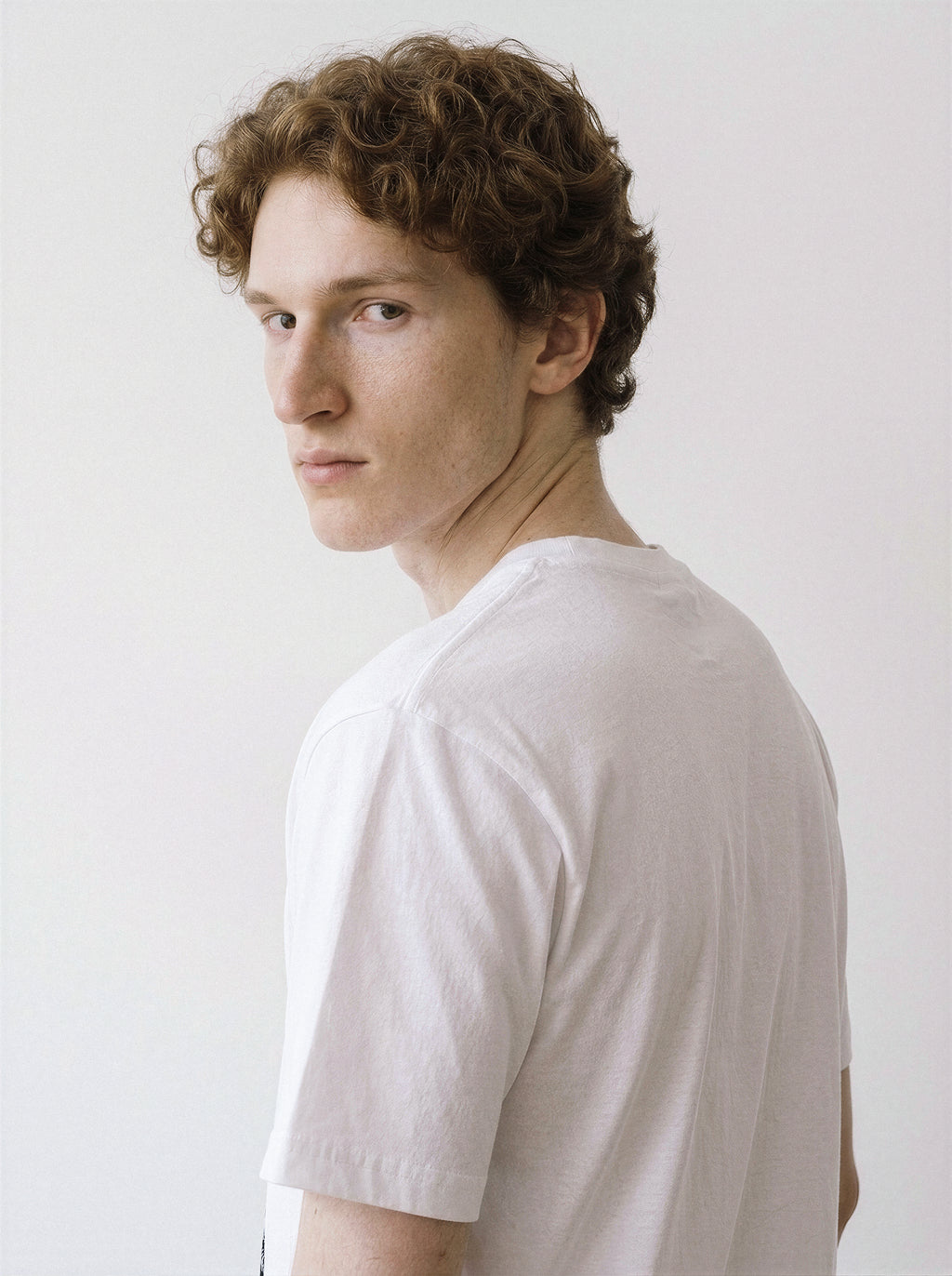 A back and shoulder view of a male model looking back wearing a white blank stare portrait line art graphic tee, demonstrating the striking minimalist streetwear style.