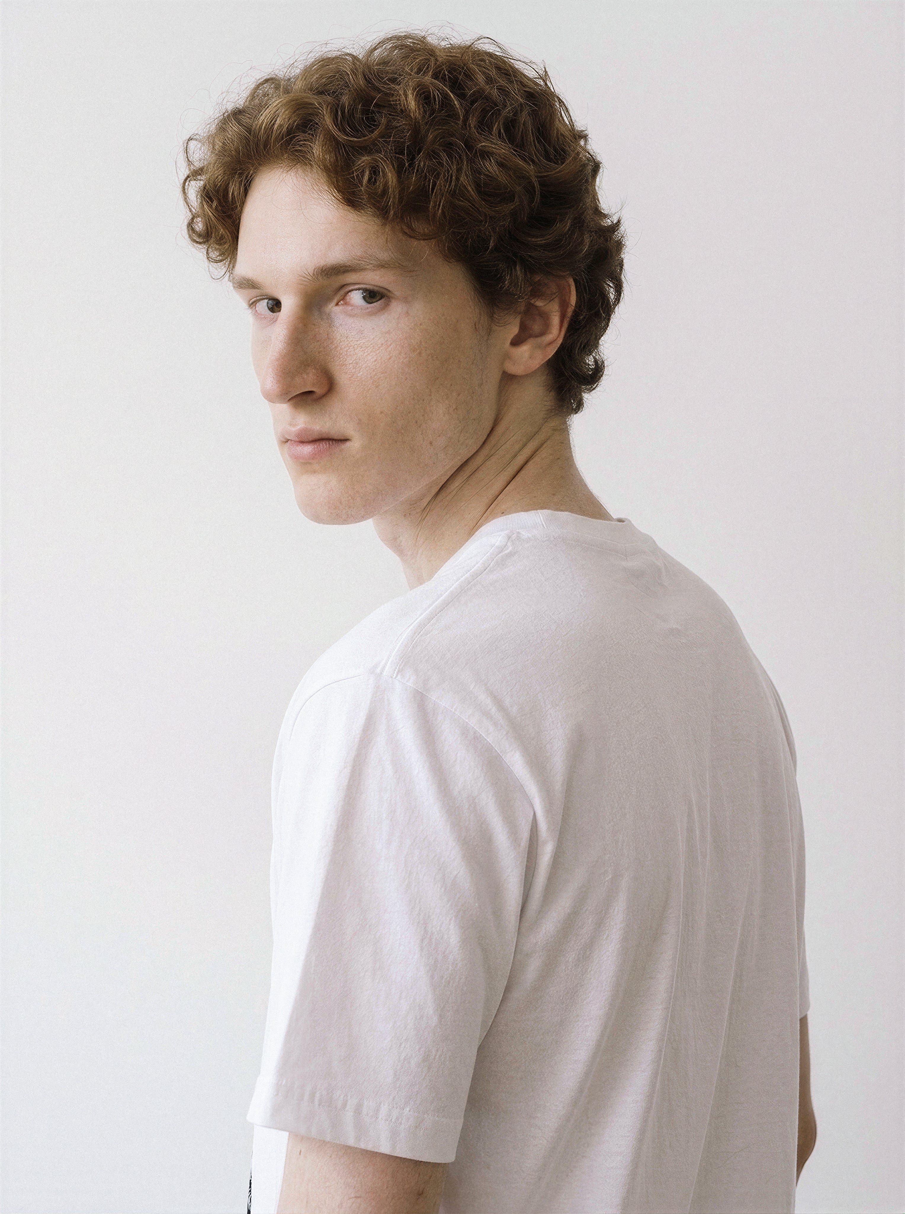 A back and shoulder view of a male model looking back wearing a white blank stare portrait line art graphic tee, demonstrating the striking minimalist streetwear style.