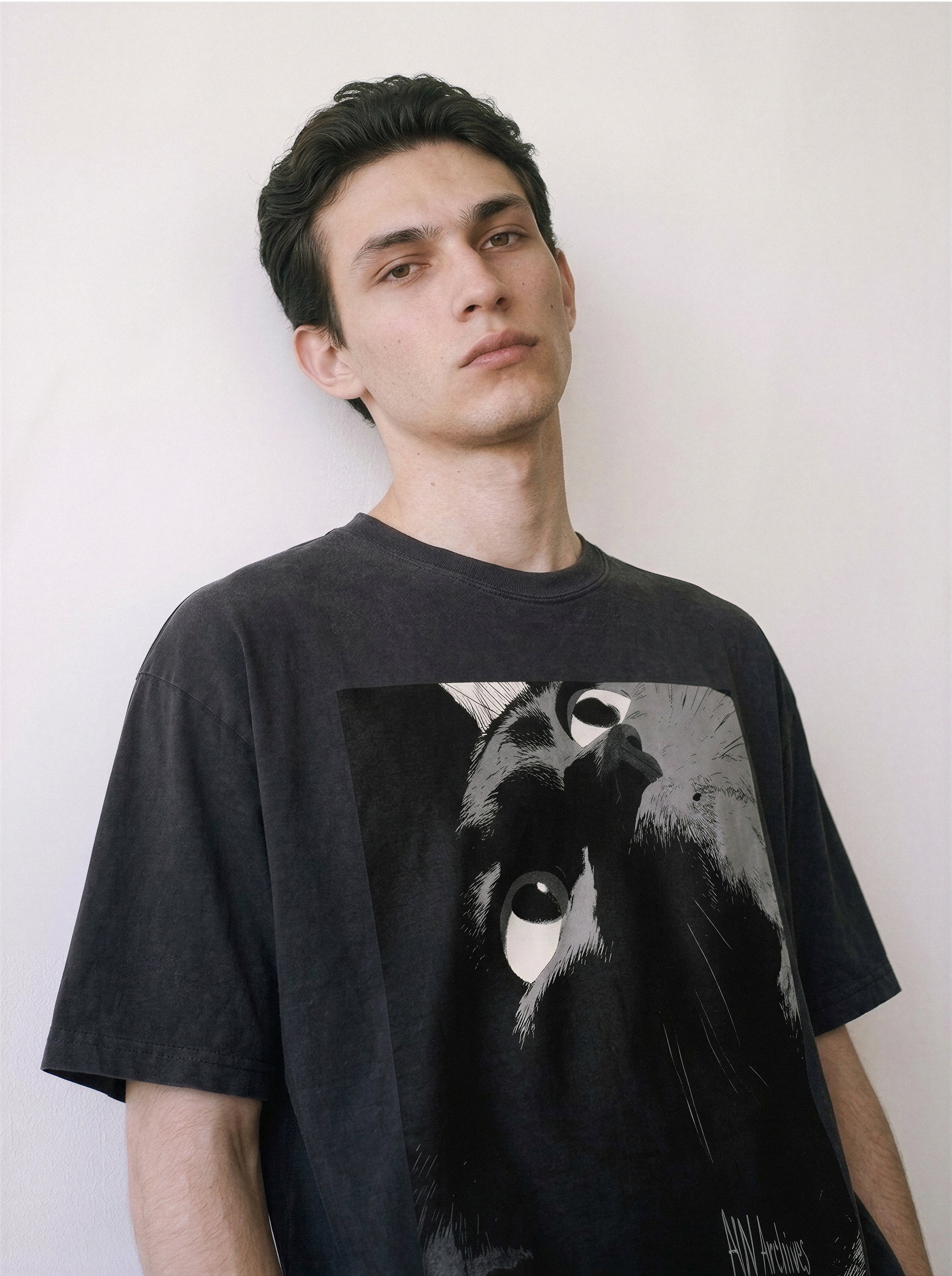 A male model leaning against a wall wearing a black cat gaze manga panel graphic tee, emphasizing the relaxed unisex fit and pure visual focus.