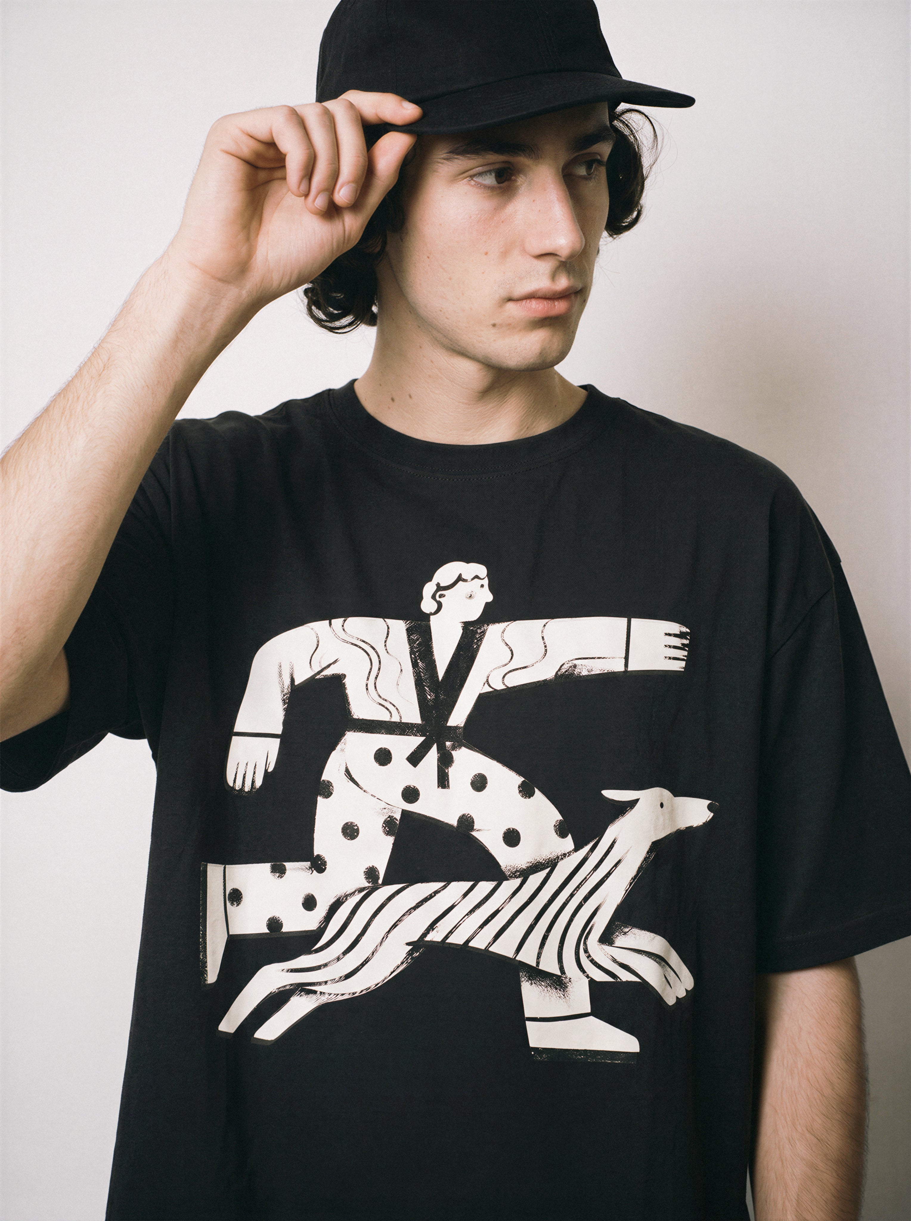 A male model adjusting his cap wearing a black confident stride boy and dog graphic tee, showcasing the uplifting positive energy streetwear vibe.