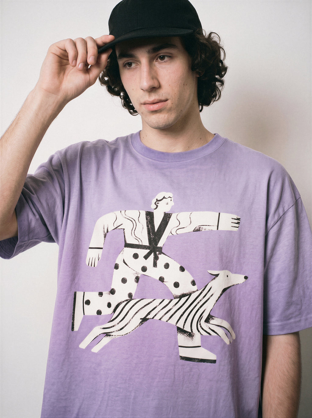A male model adjusting his cap wearing an ombre violet confident stride boy and dog graphic tee, capturing the sunny energetic illustration and good vibes apparel style.