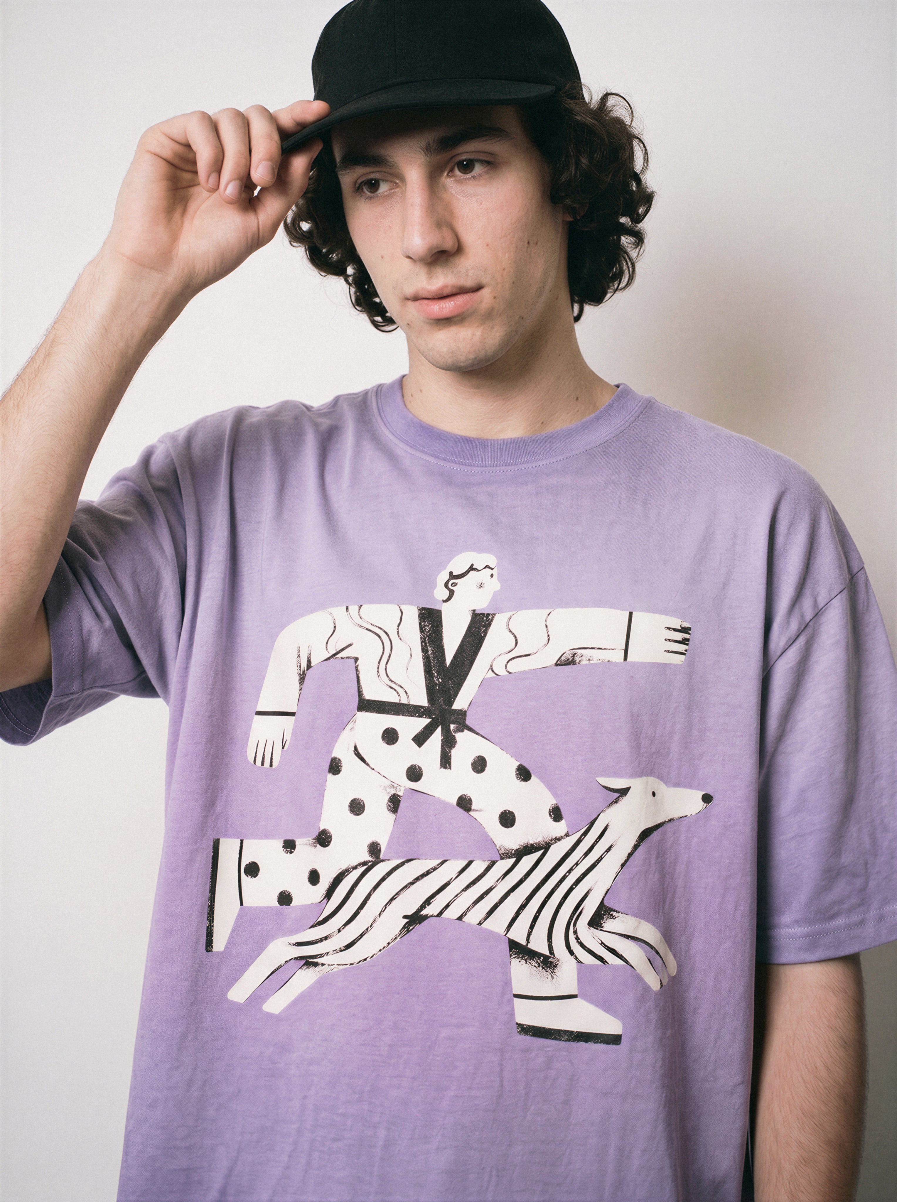 A male model adjusting his cap wearing an ombre violet confident stride boy and dog graphic tee, capturing the sunny energetic illustration and good vibes apparel style.