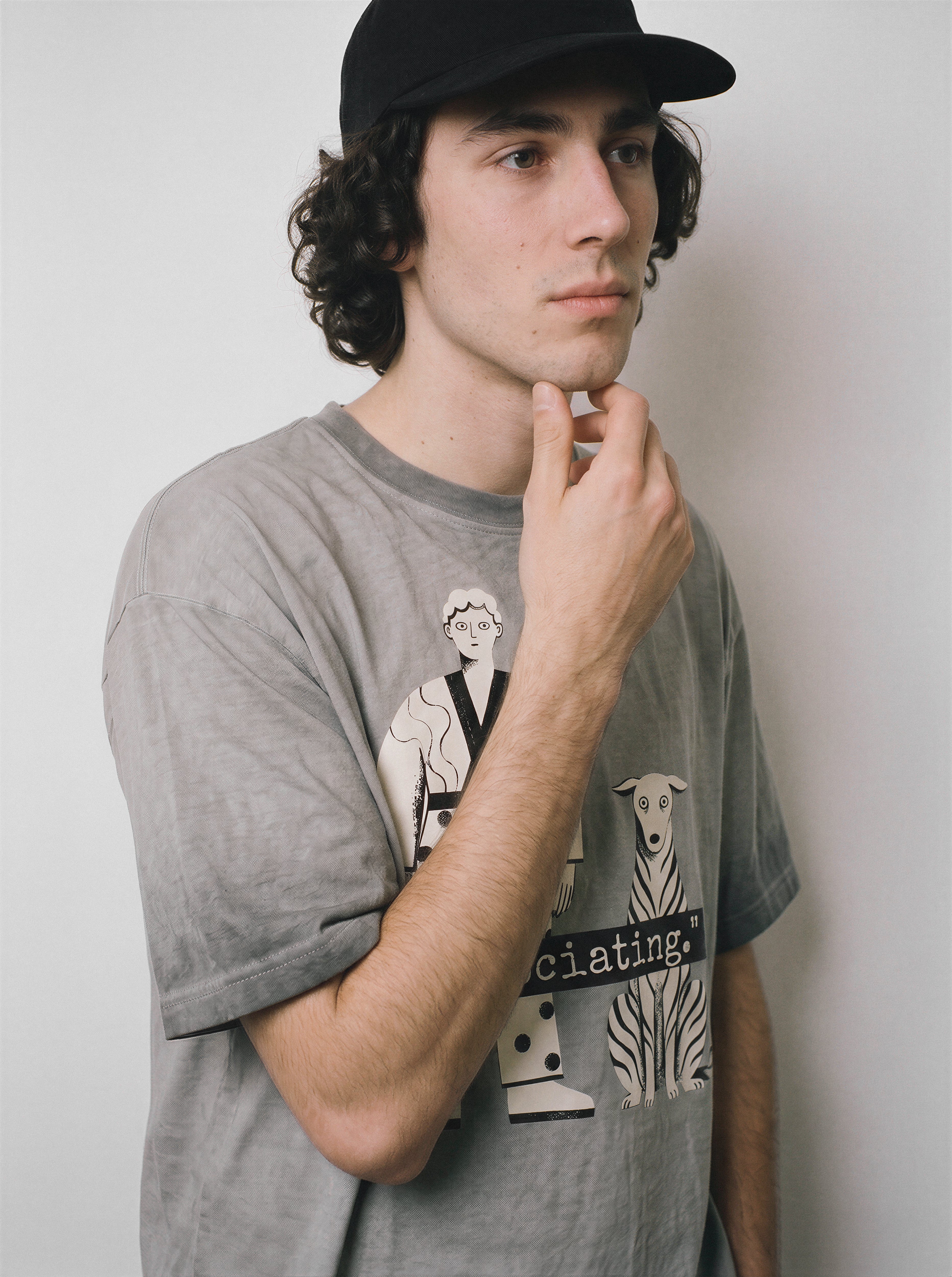 A male model resting his hand on his chin wearing an ombre grey dissociating ironic graphic tee, capturing the relatable blank stare graphic and quirky mental state aesthetic.