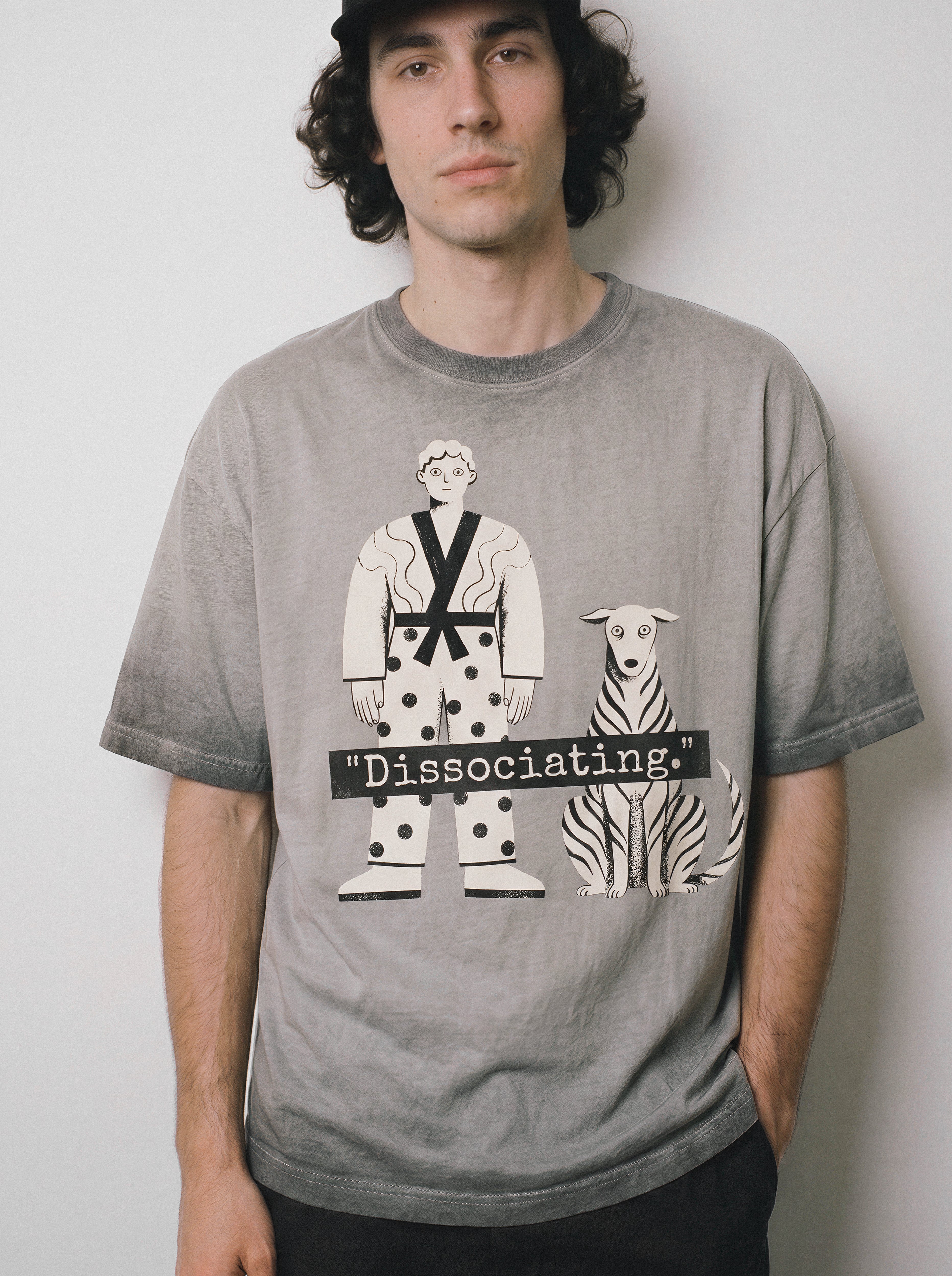 A front view of a male model with a deadpan expression wearing an ombre grey dissociating ironic graphic tee, showcasing the sarcastic boy and dog print and Gen Z existential streetwear vibe.