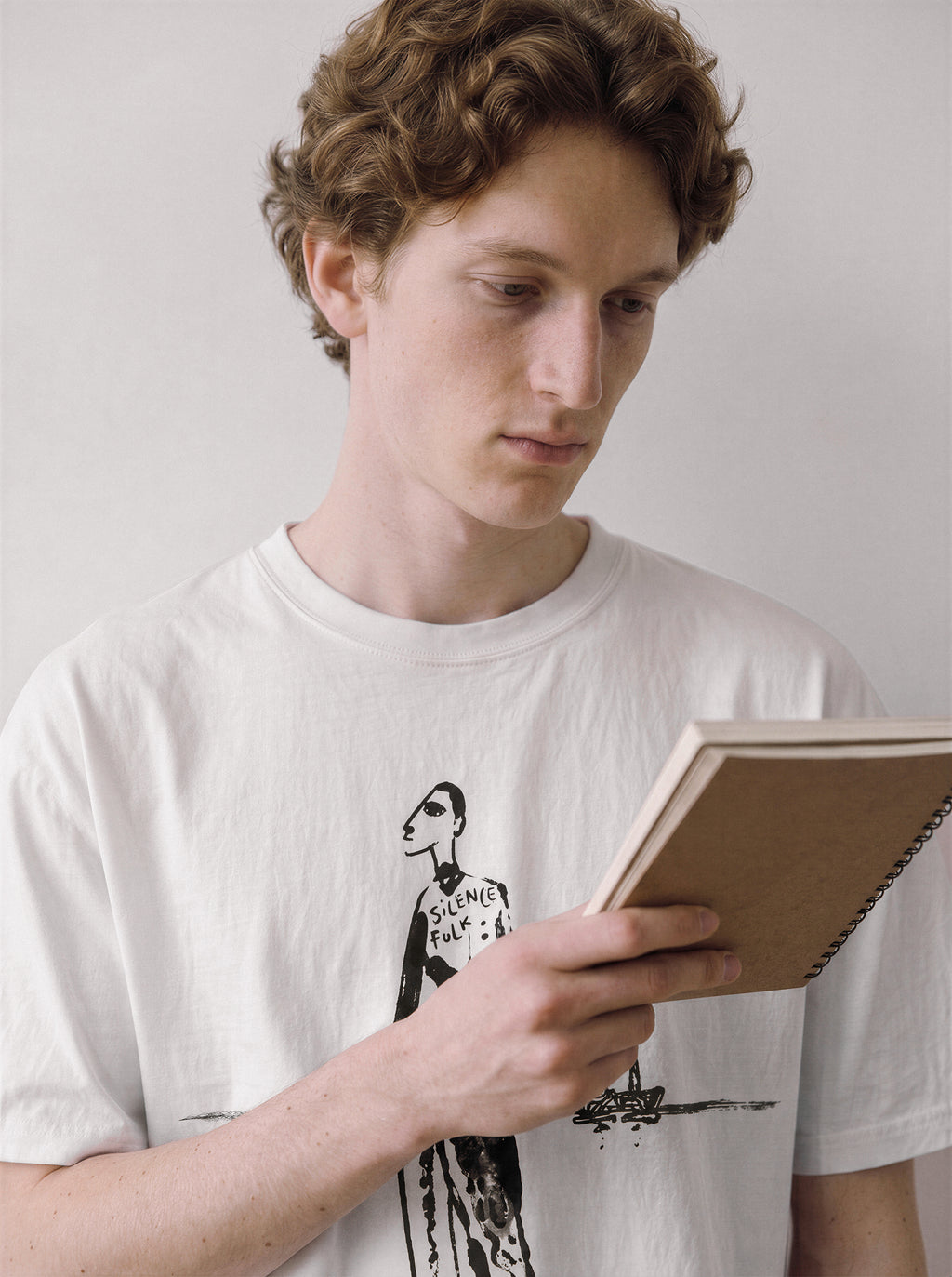 A male model looking down at a notebook wearing a white mysterious elongated figure line art graphic tee, highlighting the clean minimalist streetwear aesthetics and raw ink sketch.