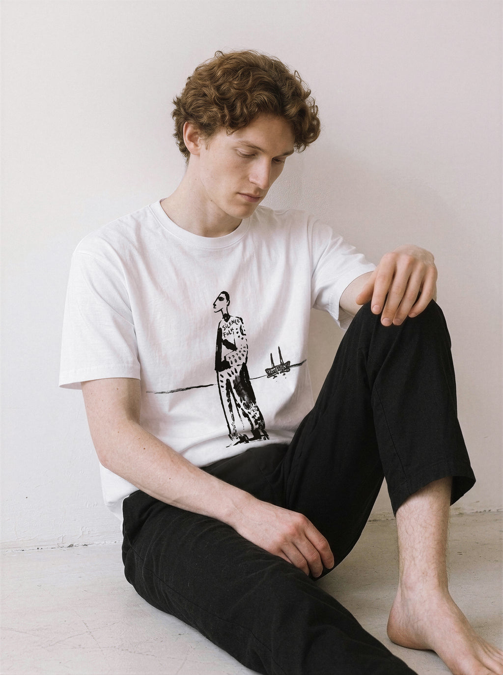 A male model sitting on the floor wearing a white mysterious elongated figure line art graphic tee, capturing a relaxed minimalist streetwear vibe with distinctive graphic details.