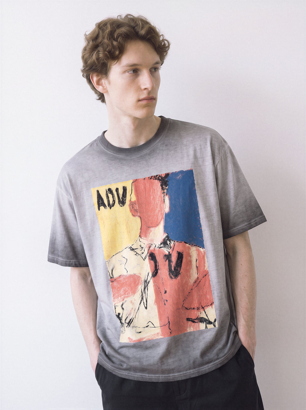 A male model with hands in his pockets wearing an ombre grey faceless portrait oil pastel graphic tee, featuring the heavy strokes and abstract art style.